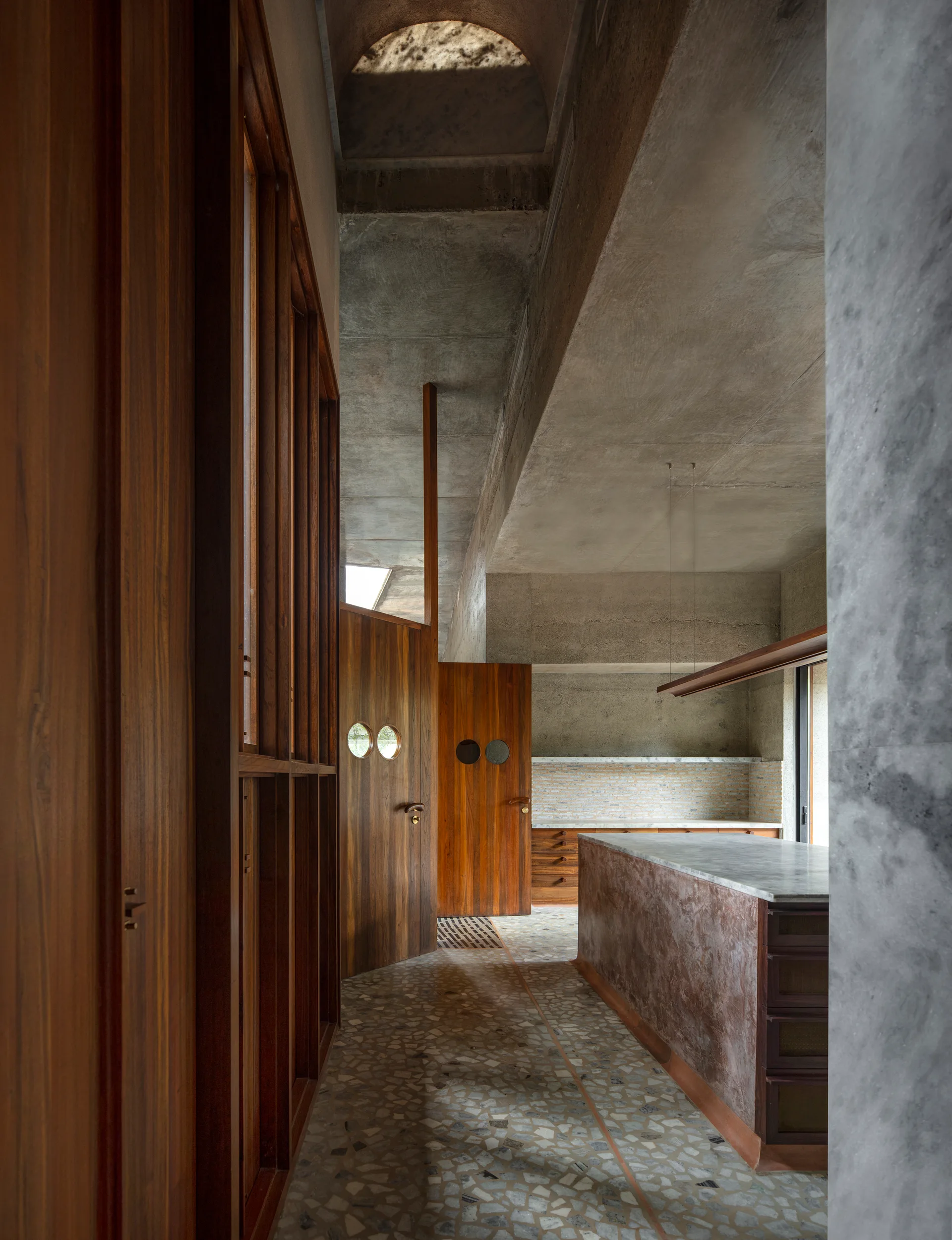 A modern kitchen features raw concrete walls, warm wood doors with circular porthole windows, terrazzo floors, and a marble-topped island.