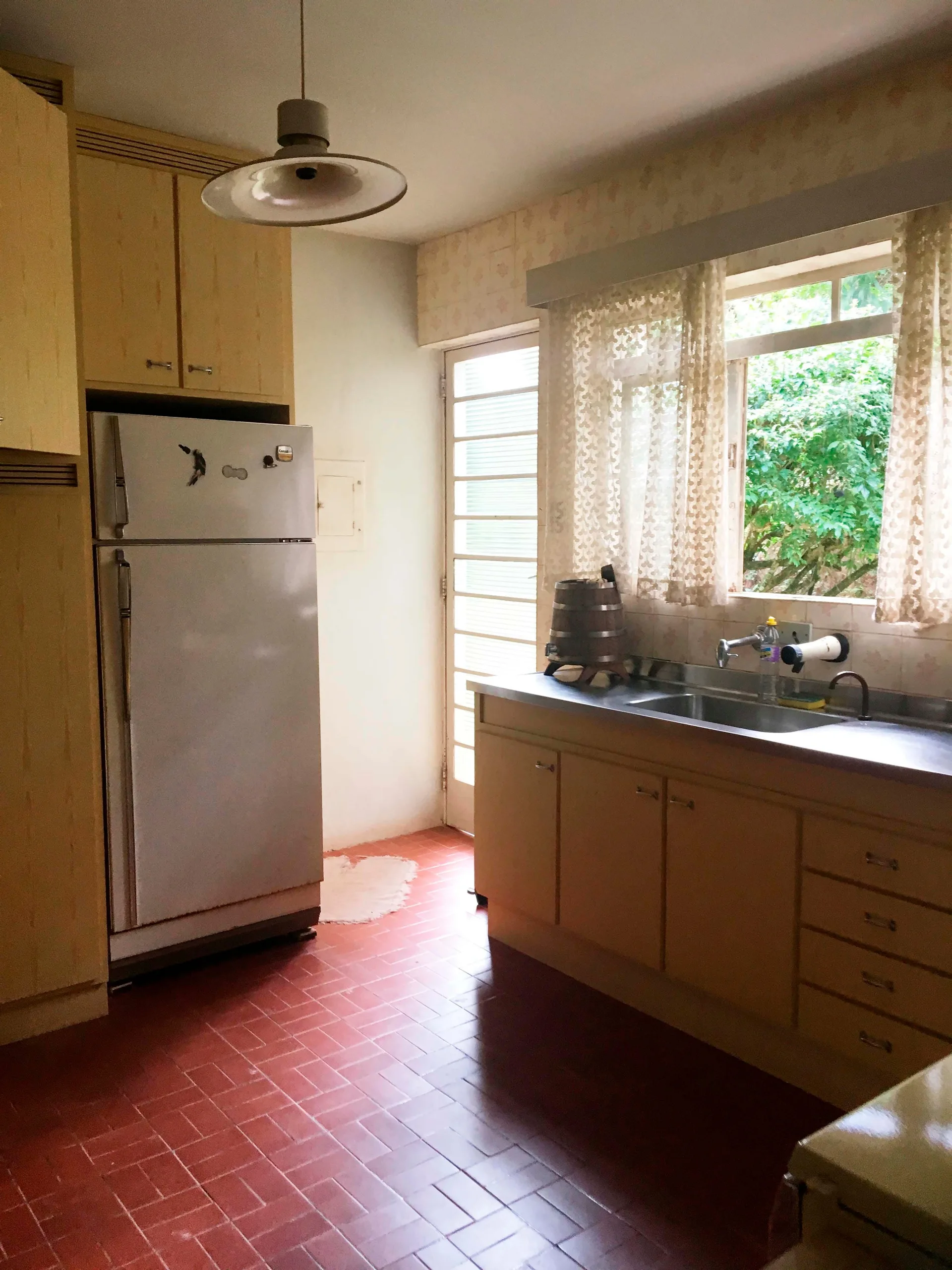 A traditional kitchen featuring light wood cabinetry, a stainless steel sink with lace curtains, and red tile flooring.