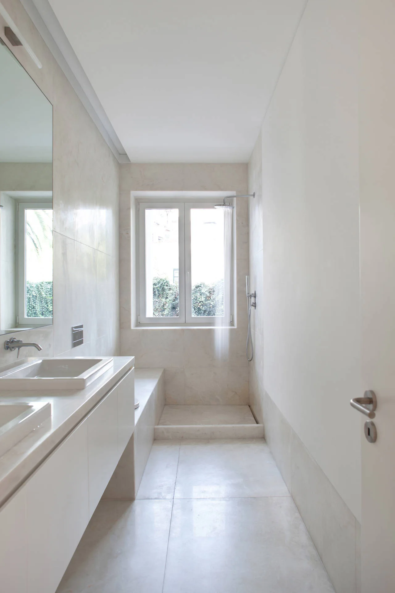 A narrow minimalist bathroom featuring a white double vanity, large-format beige stone tiles, and a window at the end of the shower enclosure.