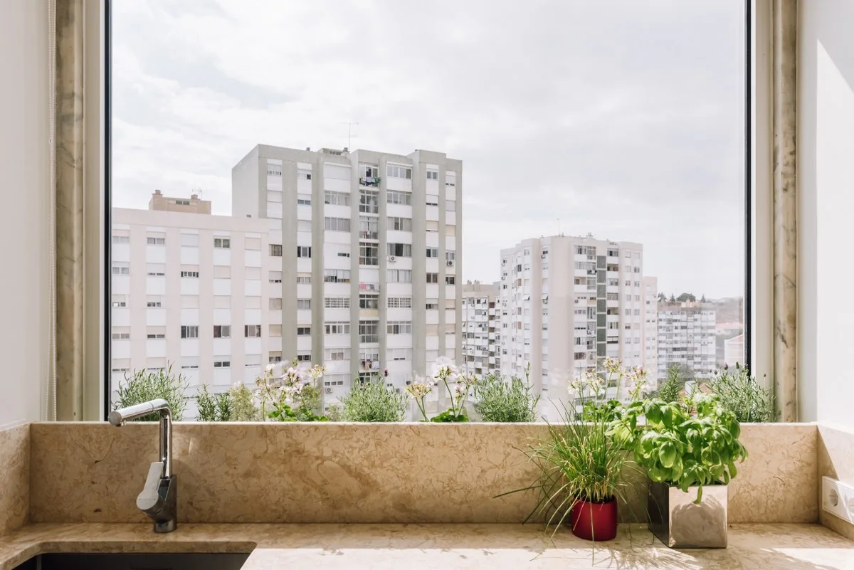 A beige veined stone countertop and low backsplash sit below a large window overlooking city buildings, styled with small potted herbs.
