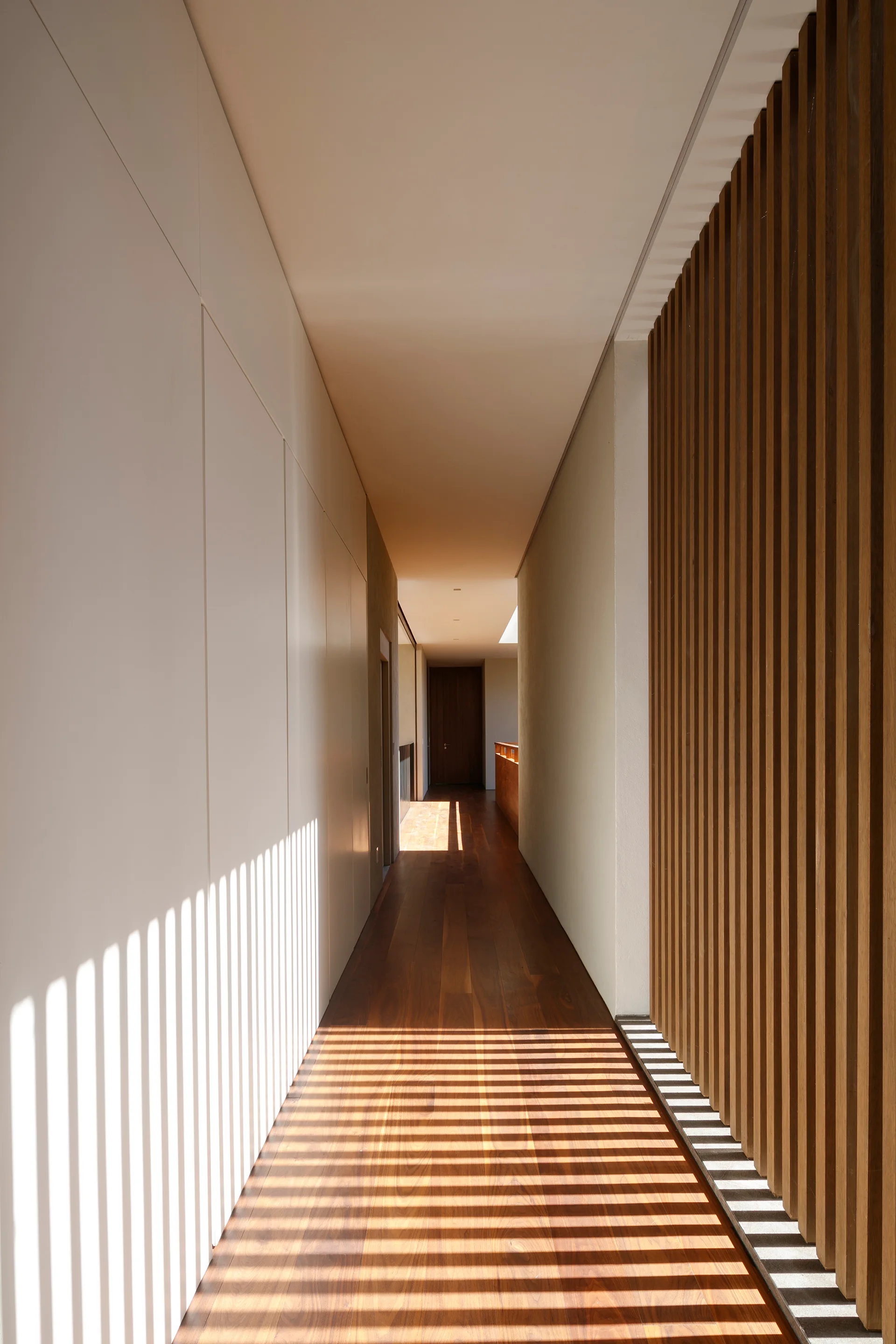 A long minimalist hallway with dark wood flooring, a smooth white wall, and a vertical wood slat screen casting linear shadows.