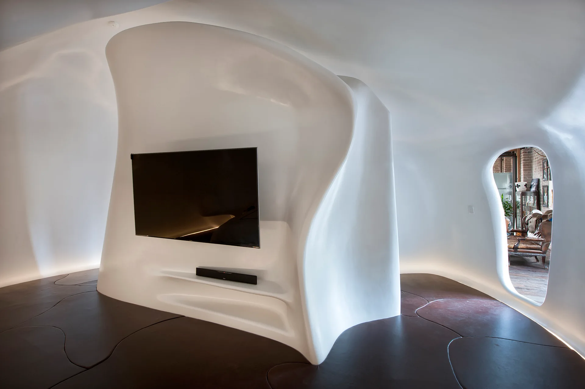 A white sculptural plaster wall with a mounted television stands on a dark brown floor next to a curved organic archway.