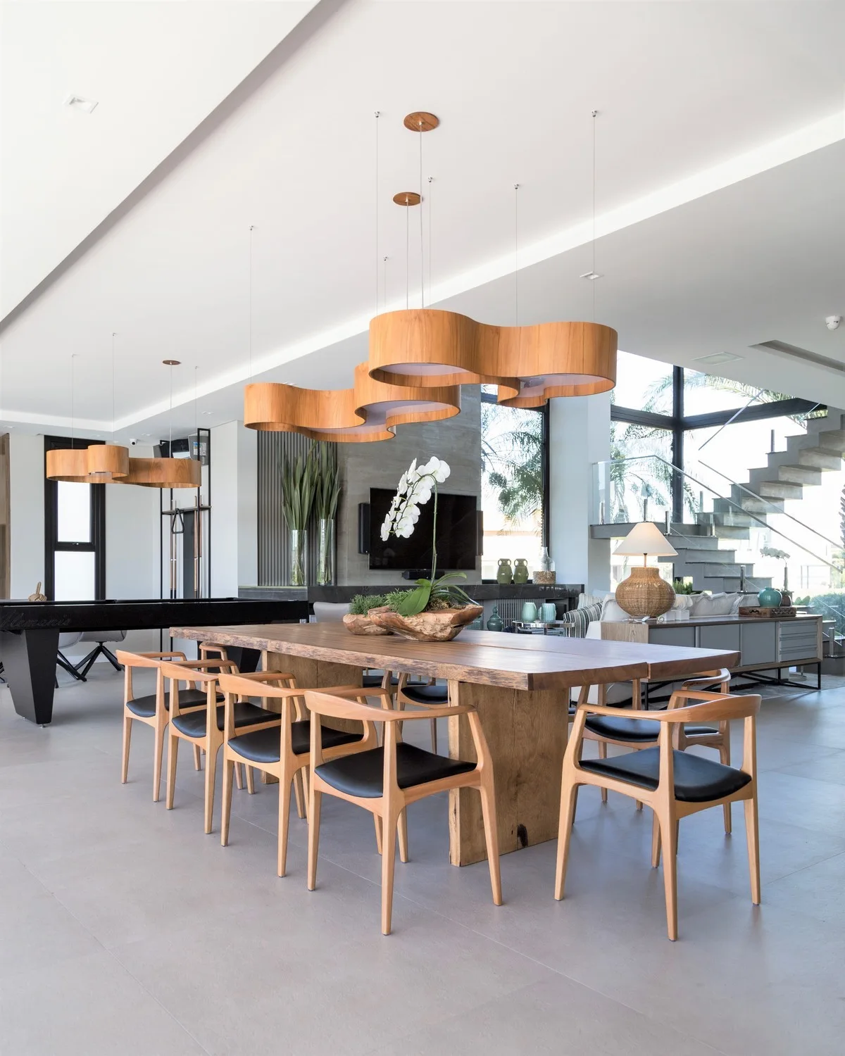 A modern dining room featuring a long live-edge wooden table, matching chairs with black seats, and wavy wood pendant lights overhead.