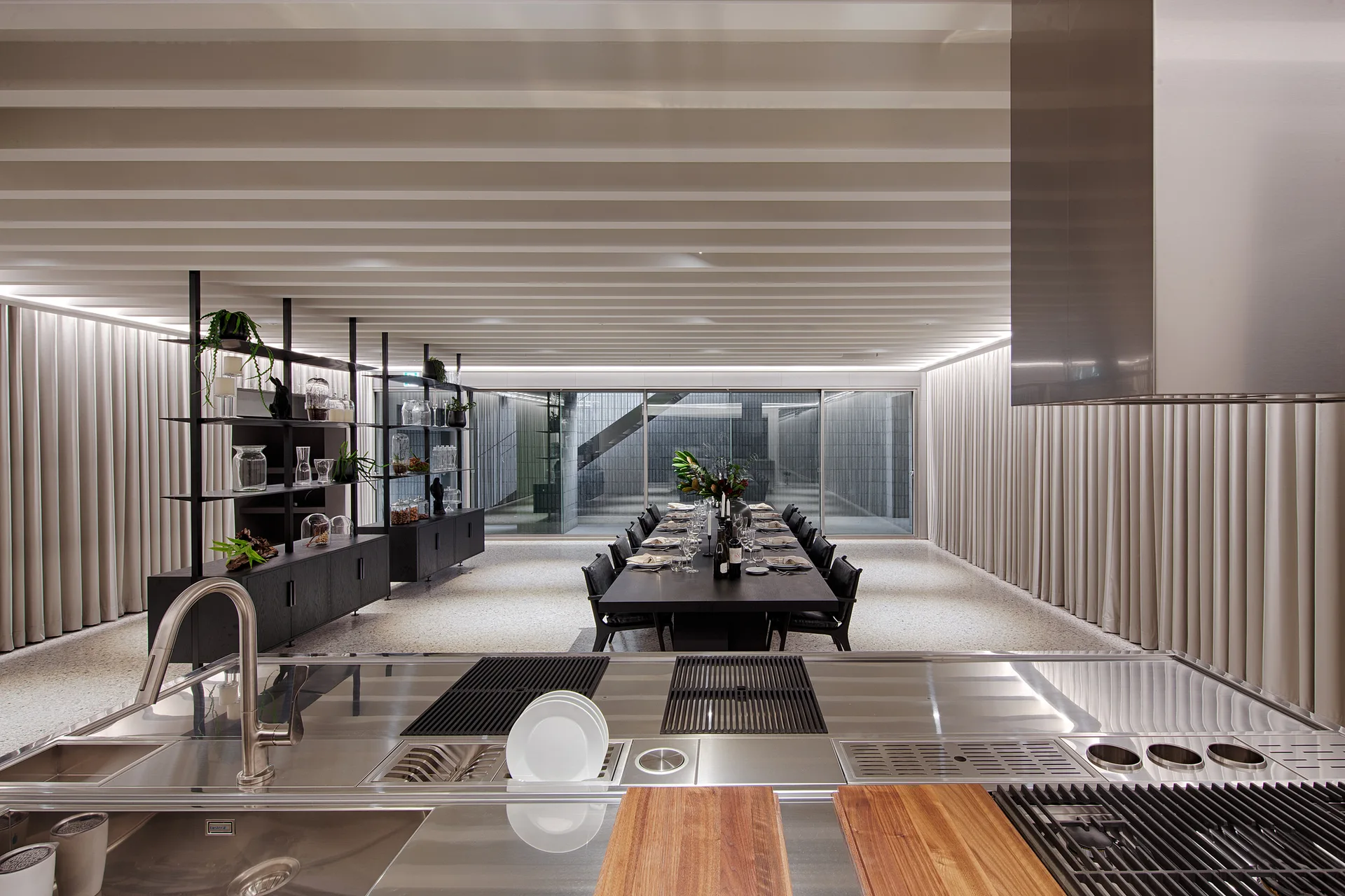 A professional stainless steel kitchen island stands in the foreground of a spacious dining room with a long black table and slatted ceiling.