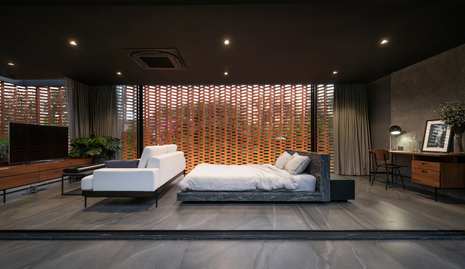 A spacious bedroom featuring a low stone platform bed, a white sofa, and a large terracotta brick lattice screen wall.