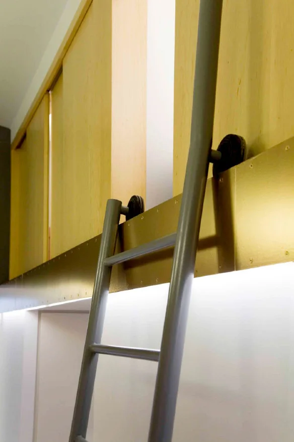 A grey metal rolling ladder hangs from a gold-toned track mounted against light wood sliding cabinet doors.
