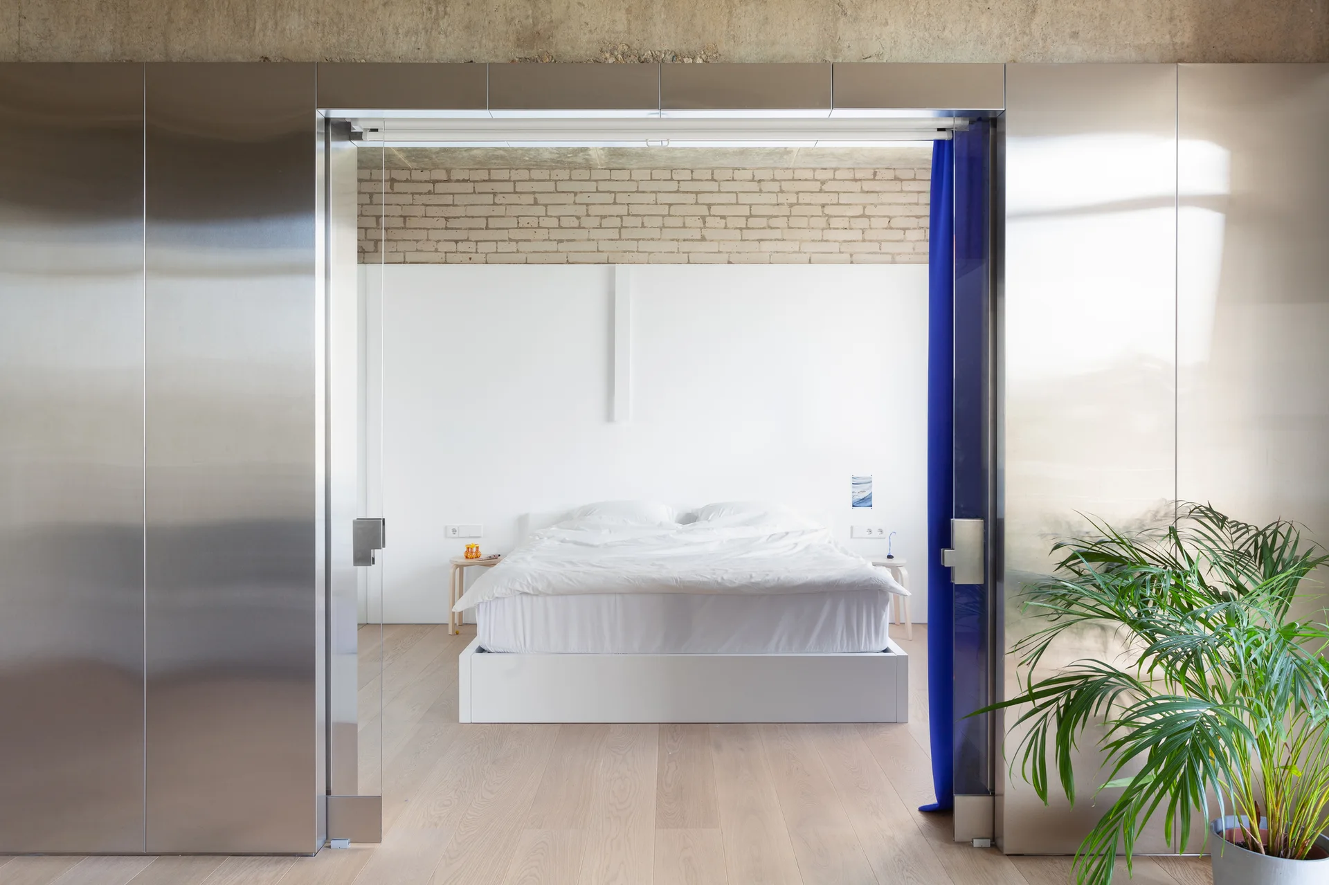 A minimalist bedroom with a white bed is viewed through a wide doorway framed by brushed stainless steel wall panels.