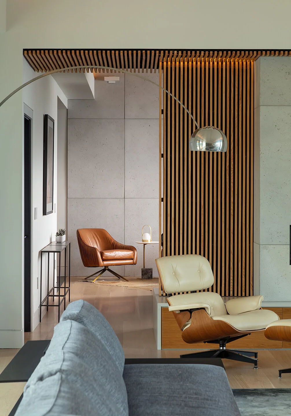 An open-plan living area with an off-white lounge chair, a vertical wood slat partition, concrete panel walls, and a tan leather armchair.