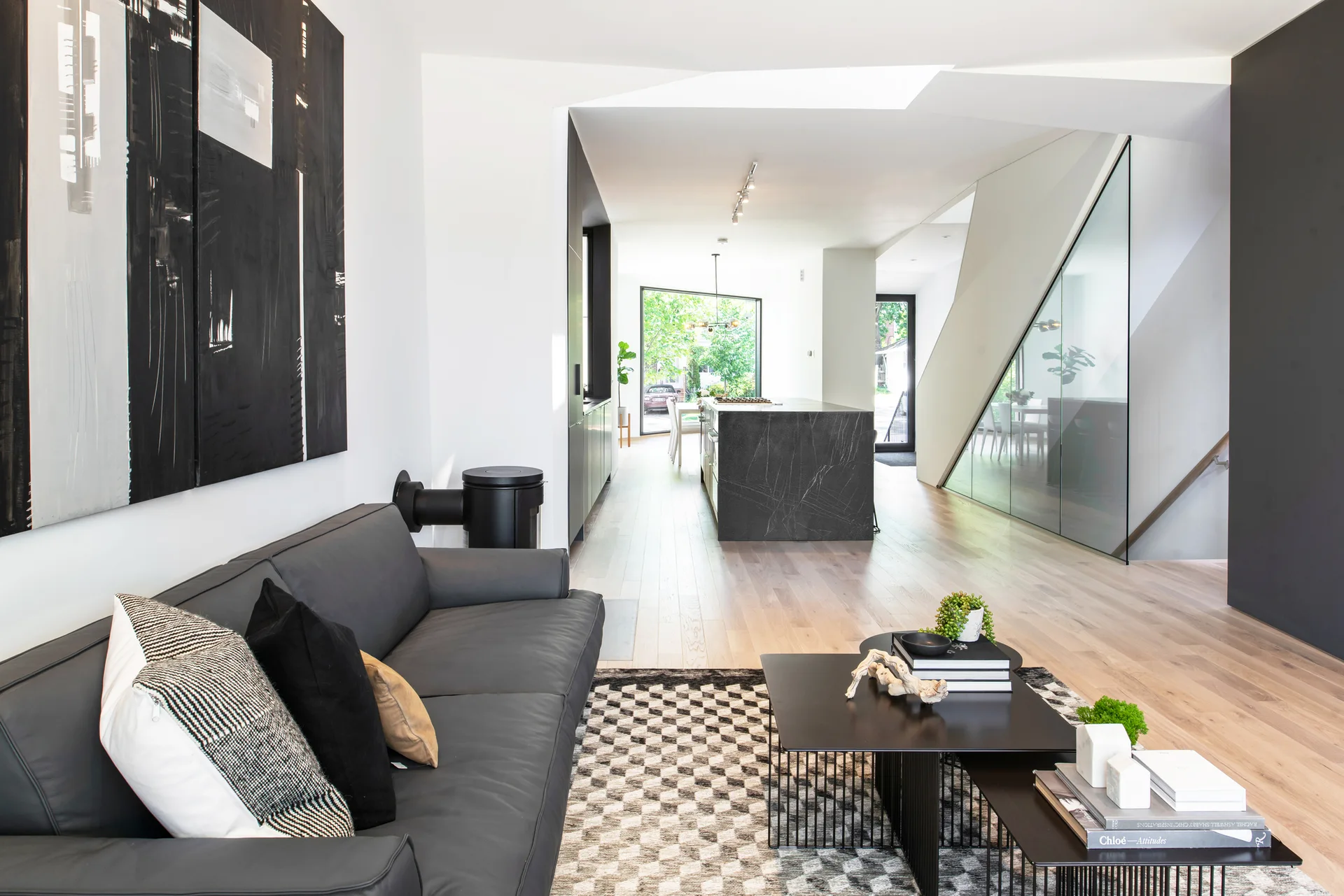 A minimalist living room with a black leather sofa and a geometric rug leads to an open kitchen with a dark stone island and an angled glass staircase.