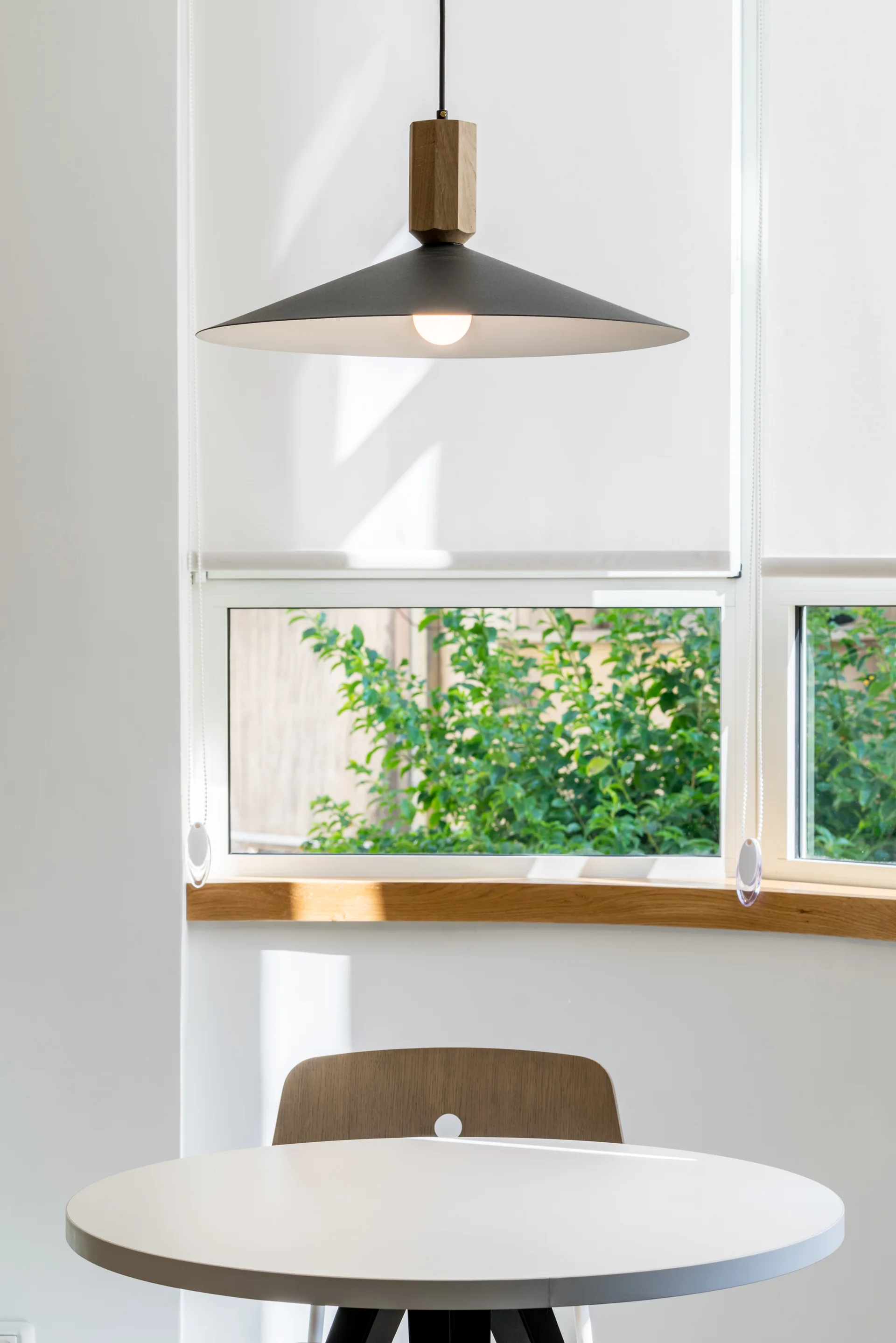 A minimalist dining nook features a round white table, a wooden chair, and a black cone-shaped pendant light in front of a window with green foliage.