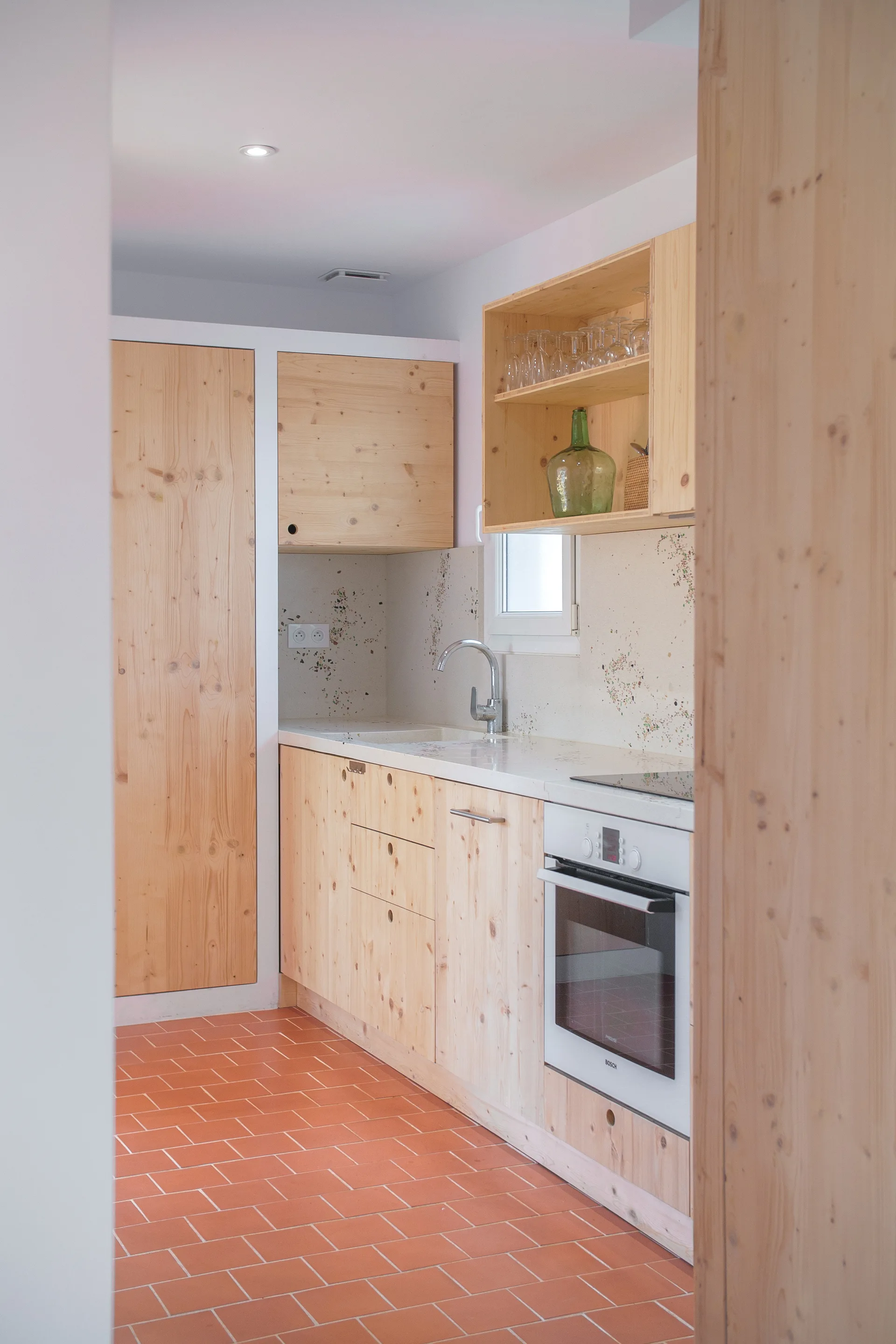A small galley kitchen features knotty light wood cabinets, a speckled backsplash, a white oven, and terracotta tiled floors.