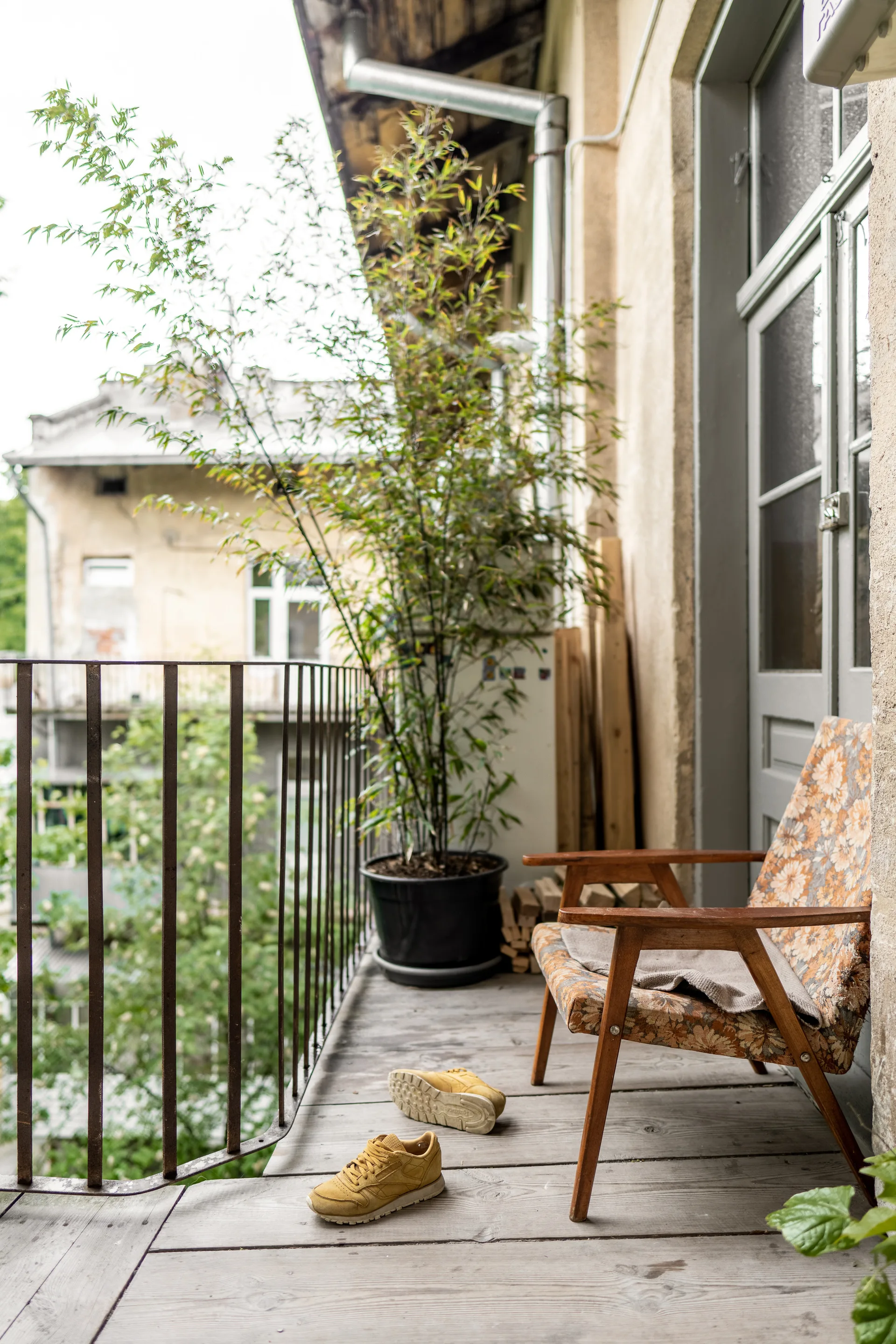 A weathered wood-plank balcony features a vintage floral armchair, a tall potted plant, and a metal railing.