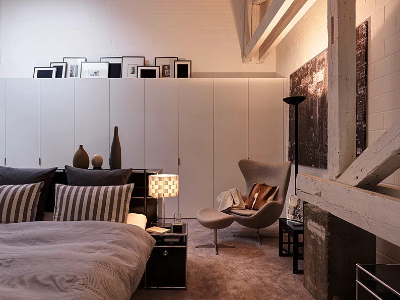 A minimalist bedroom featuring a bed with striped bedding, a grey lounge chair with ottoman, white built-in wardrobes, and exposed wooden beams.