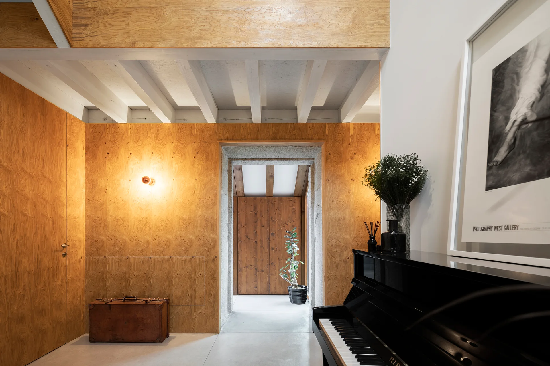 A modern hallway features warm burl wood wall paneling, white exposed ceiling beams, a black upright piano, and a stone-trimmed doorway.