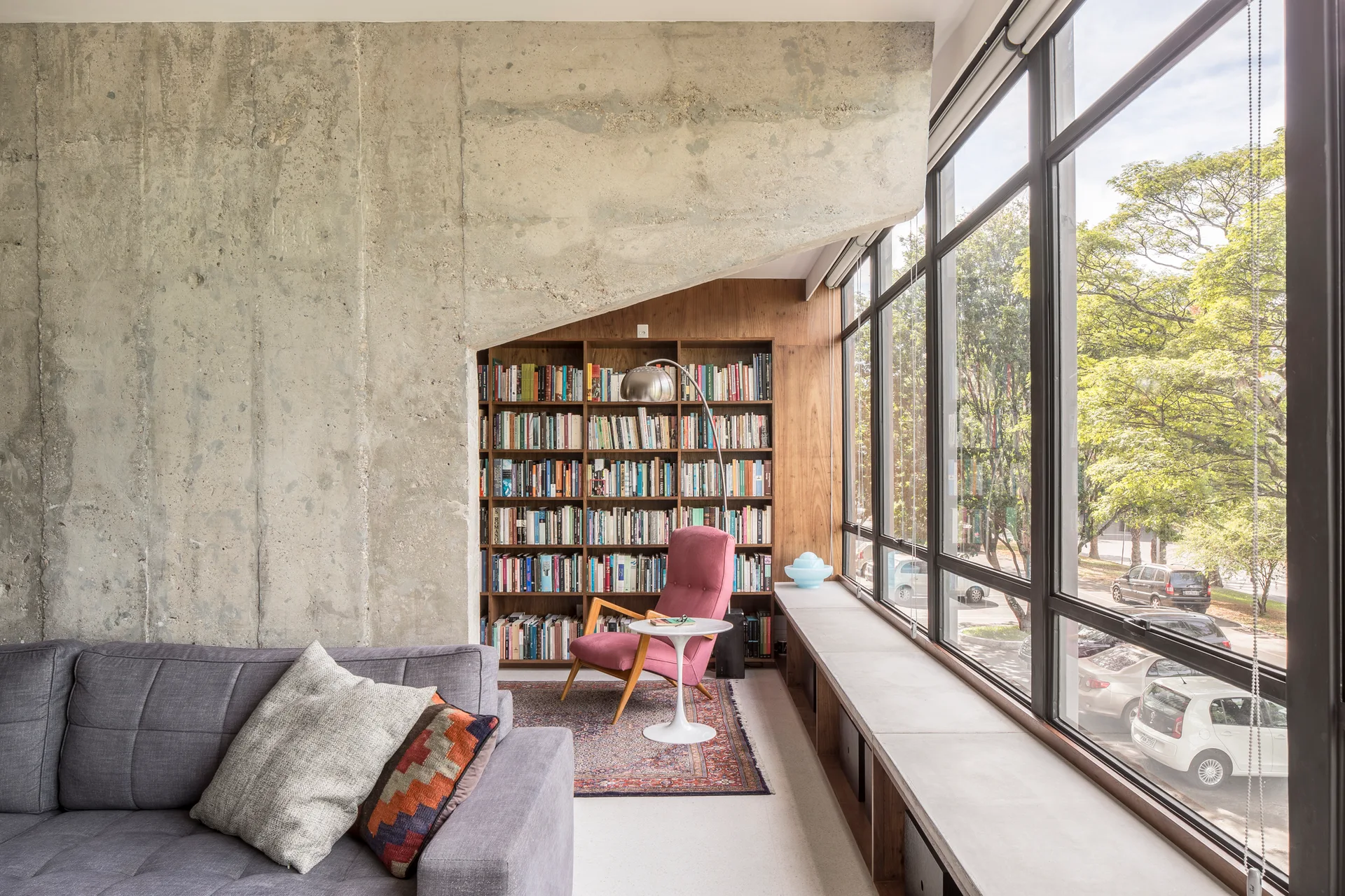 A modern living area with a raw concrete wall, built-in wooden bookcase, pink armchair, and large windows with a concrete bench.