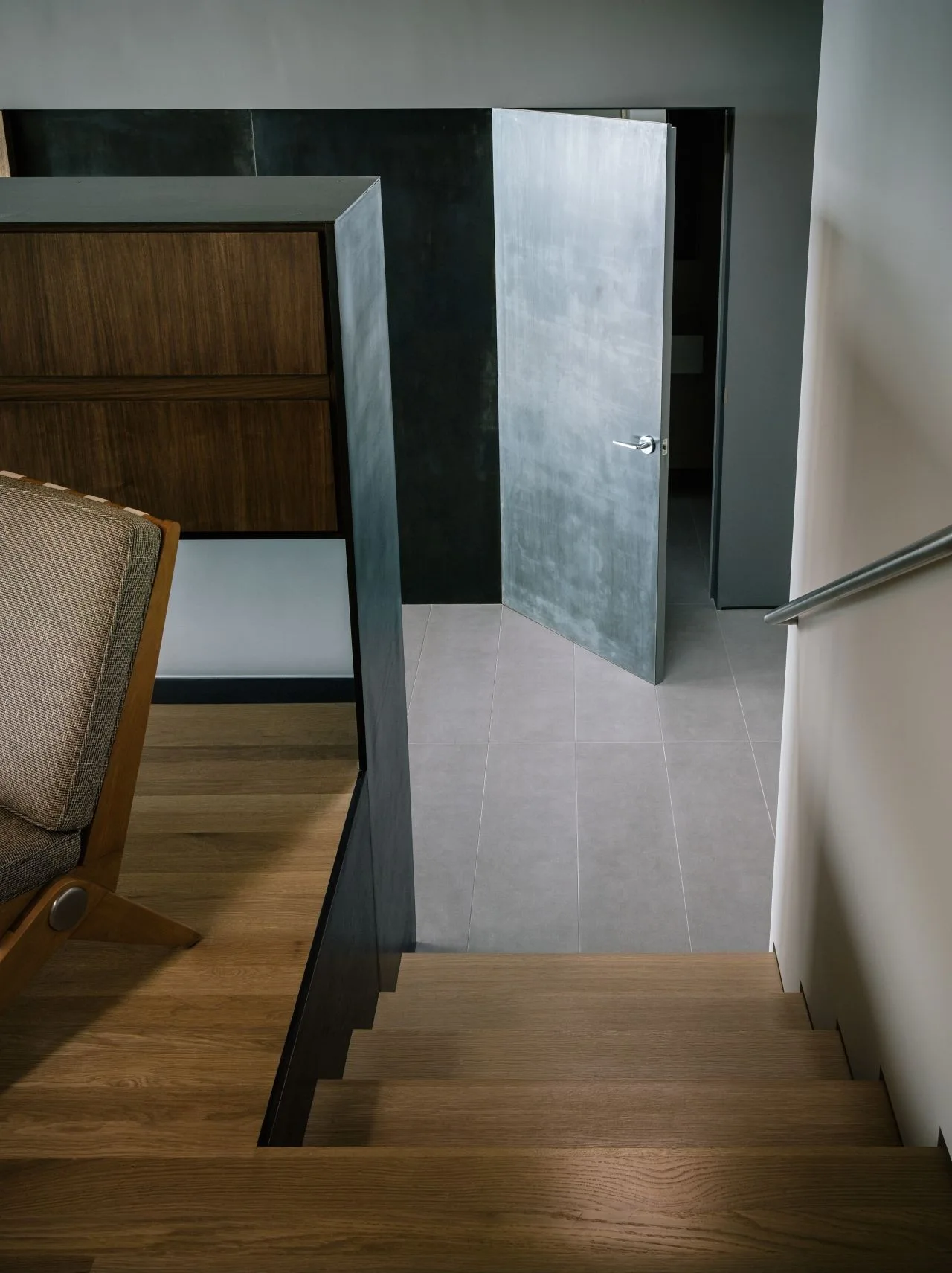 A wooden staircase descends to a grey tile landing featuring a metallic open door, a black accent wall, and a metal handrail.