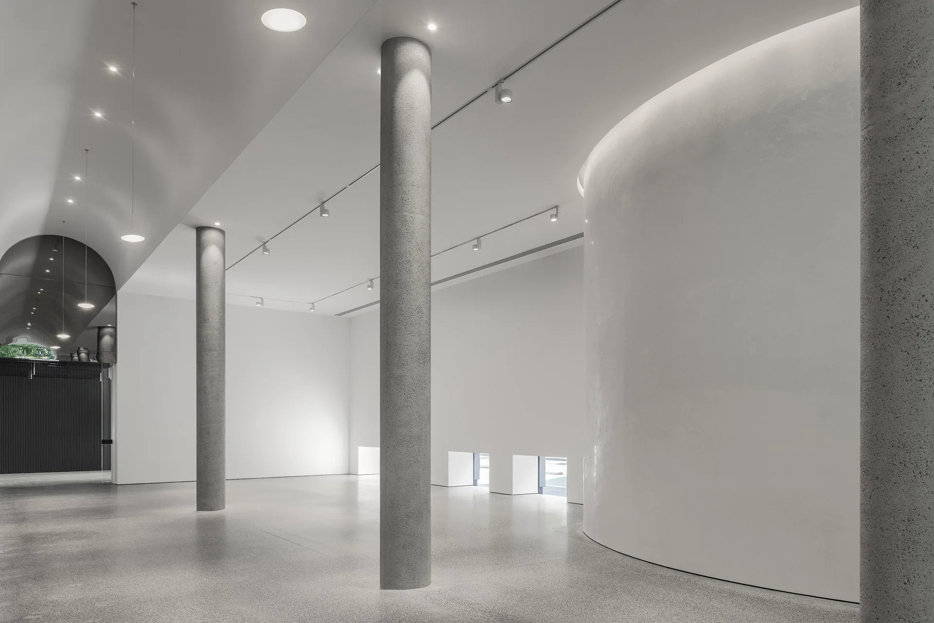 A minimalist architectural space featuring tall round concrete columns, polished grey flooring, a large curved white wall, and ceiling track lights.