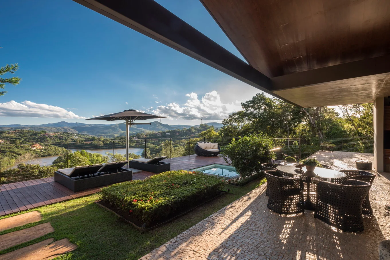 A modern outdoor patio features a stone dining terrace under a wood ceiling overhang next to a wooden deck with lounge chairs and a sunken pool.