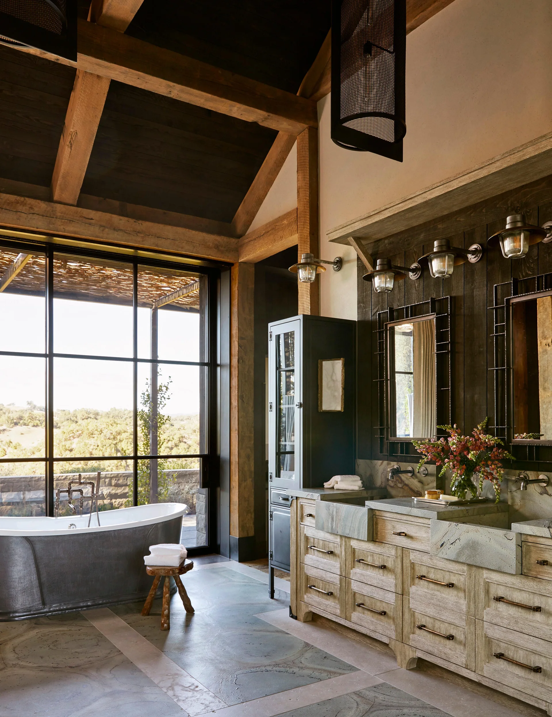 A rustic bathroom features a freestanding metal bathtub near a large window, a double wood vanity with stone sinks, and exposed timber ceiling beams.
