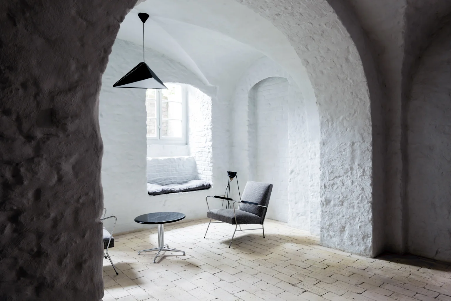 A minimalist lounge area within a vaulted white brick room featuring a grey armchair, a black side table, and a geometric black pendant light.