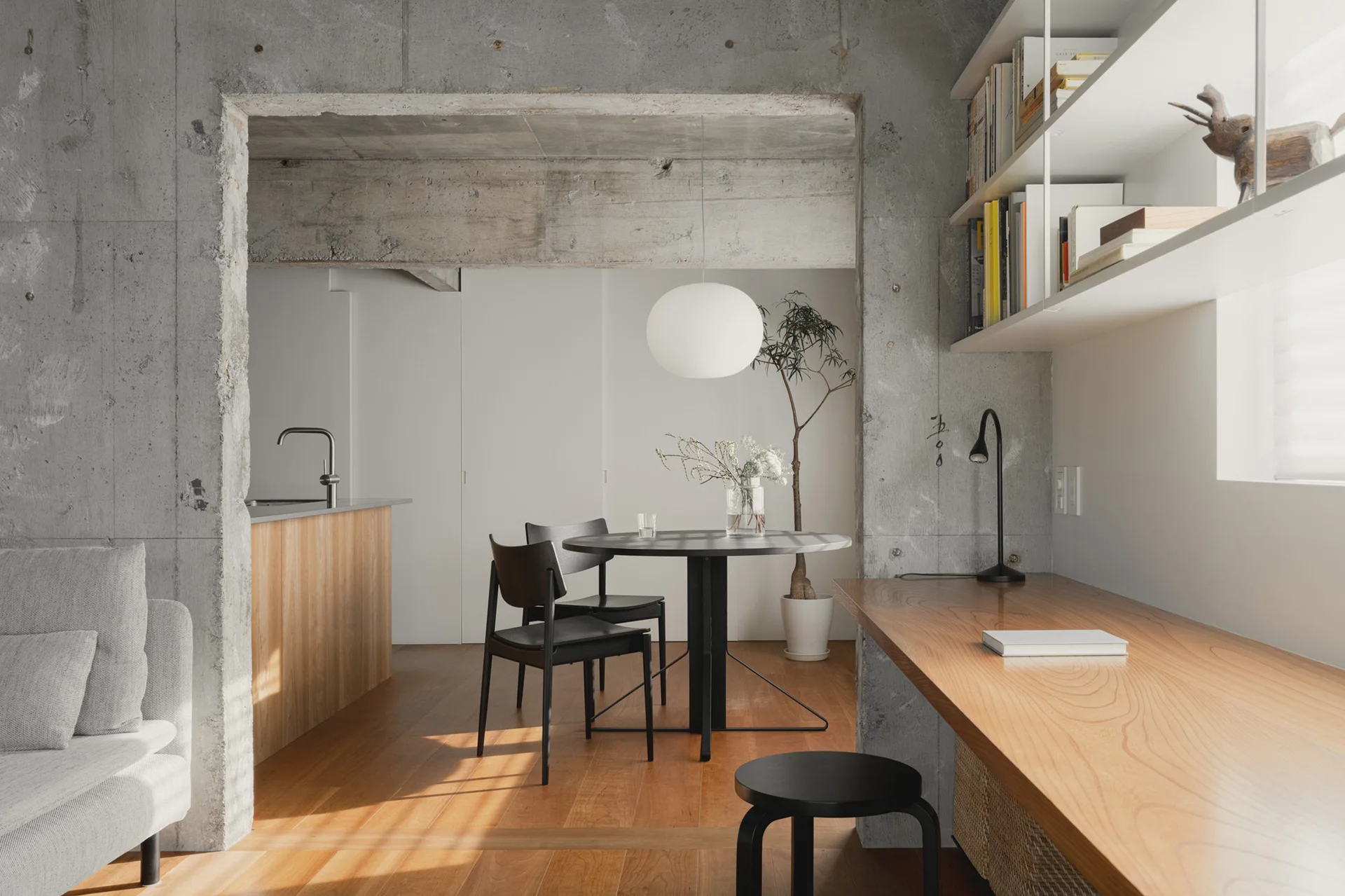 A dining area with a round black table and chairs set against raw concrete walls, featuring a long built-in wooden desk along the side.