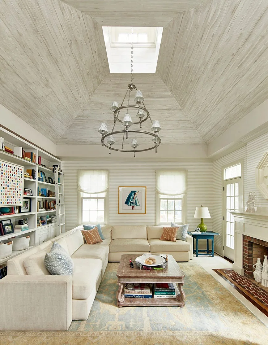 A bright living room features a cream sectional sofa, built-in bookshelves, and a vaulted wood ceiling with a central skylight and chandelier.