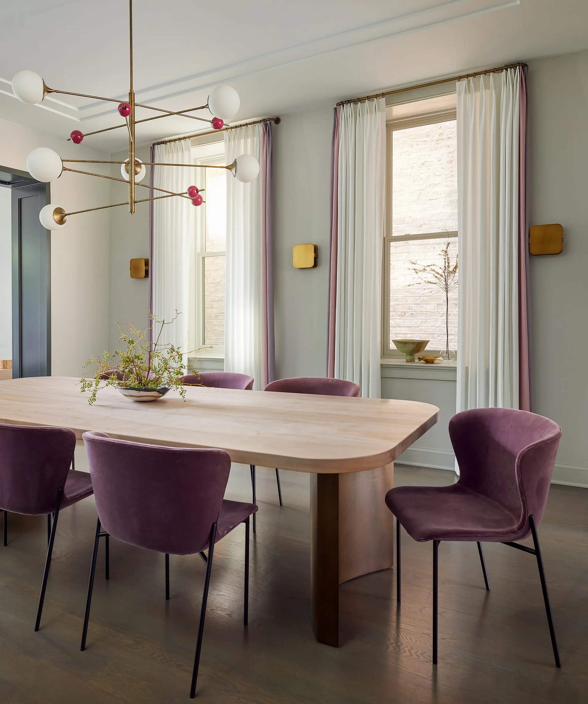 A modern dining room featuring a light wood table, plum velvet chairs, a branching brass chandelier, and two windows with white and purple curtains.