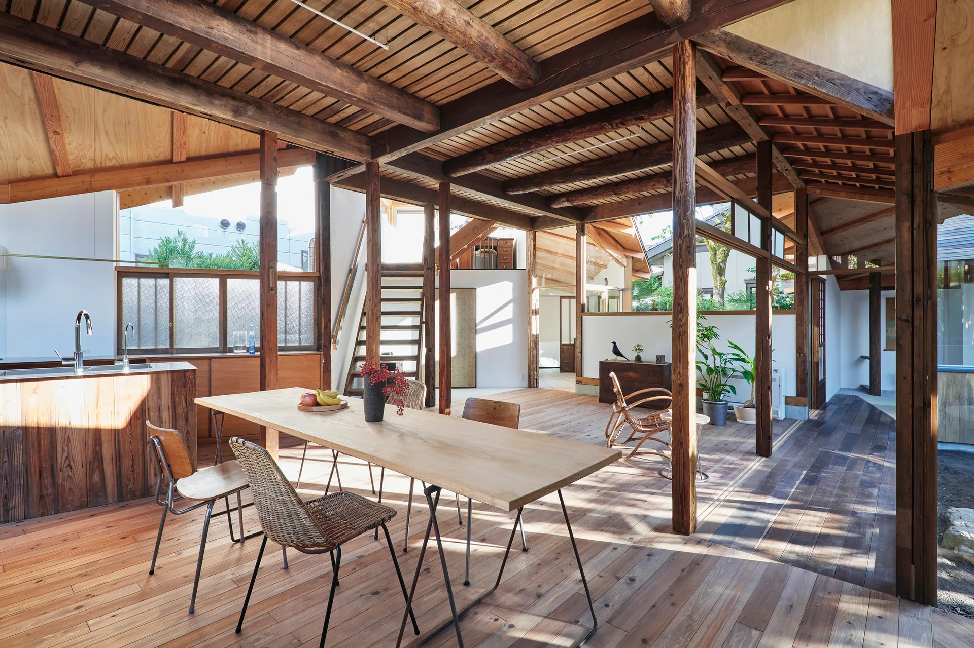 A rustic dining room features a long wooden table and chairs beneath an exposed timber ceiling with dark posts and beams.