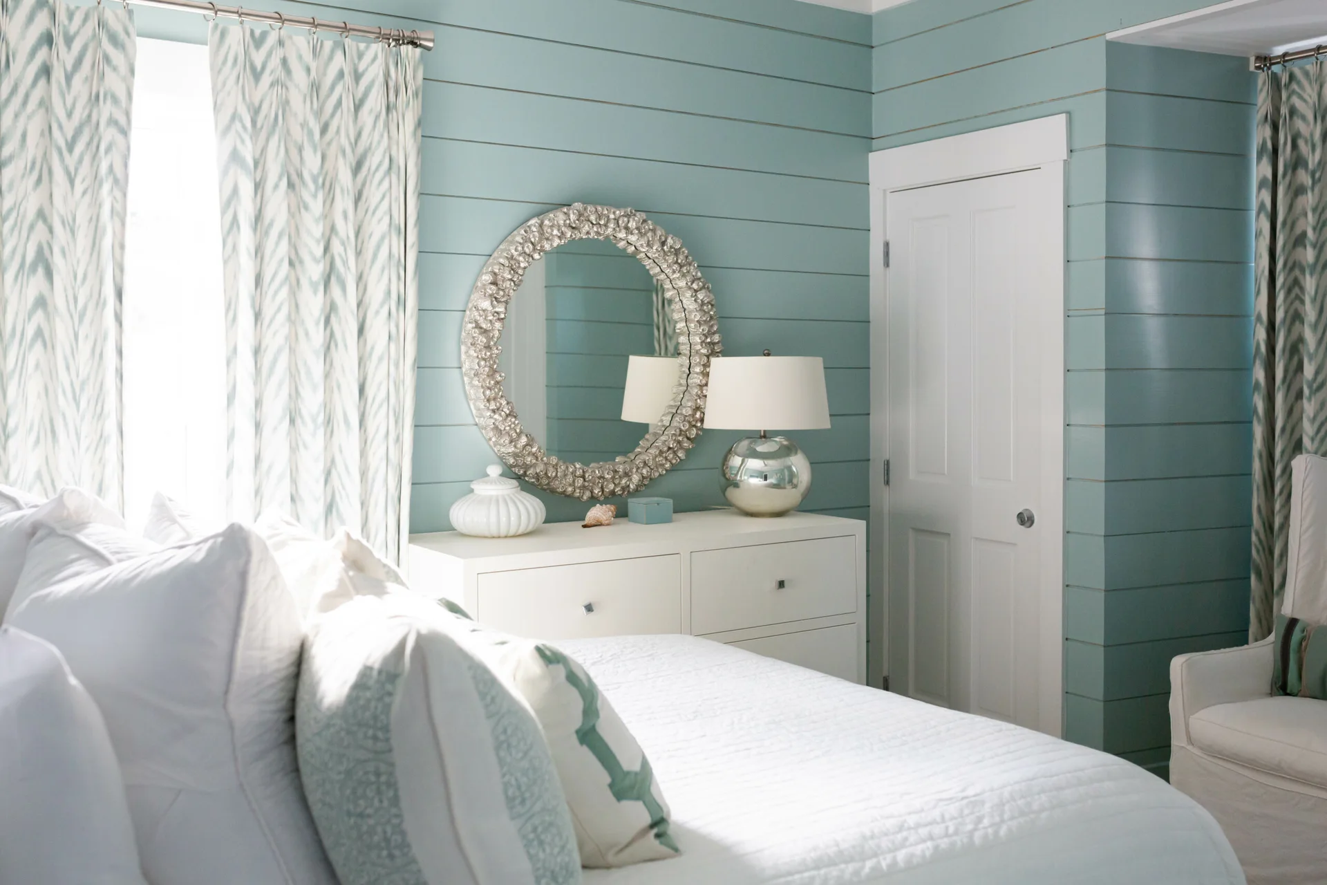 A bedroom features pale blue horizontal shiplap walls, a white dresser with a round textured mirror, a silver table lamp, and chevron patterned curtains.