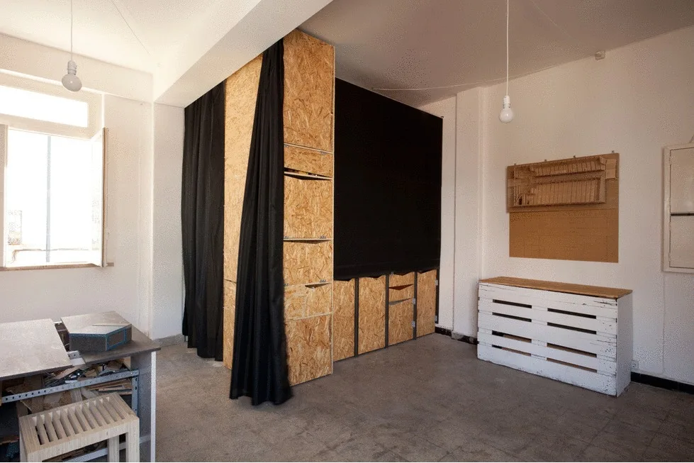 An industrial studio space featuring a large OSB storage cabinet with a black curtain, a white pallet console, and hanging bare-bulb pendant lights.