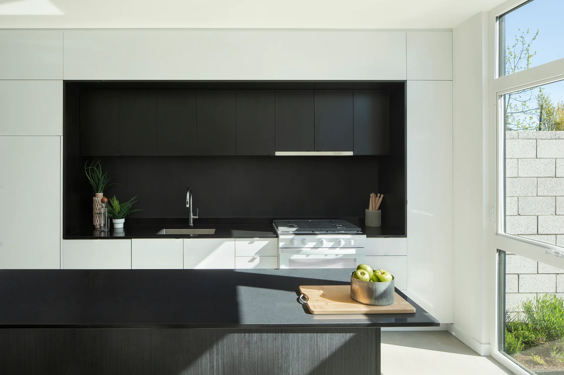 A modern kitchen features black recessed upper cabinets and countertops surrounded by flush white cabinetry next to a large window.