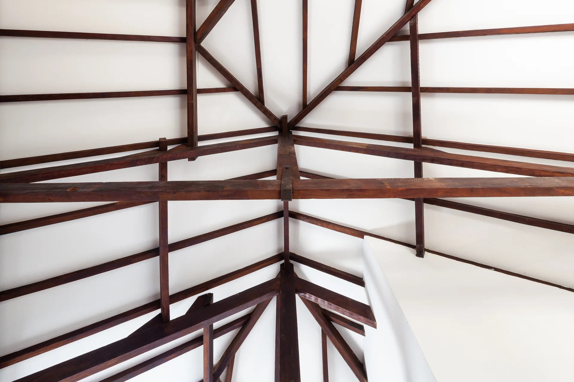 An upward shot of a vaulted ceiling featuring an intricate framework of dark brown wooden timber beams against a clean white surface.