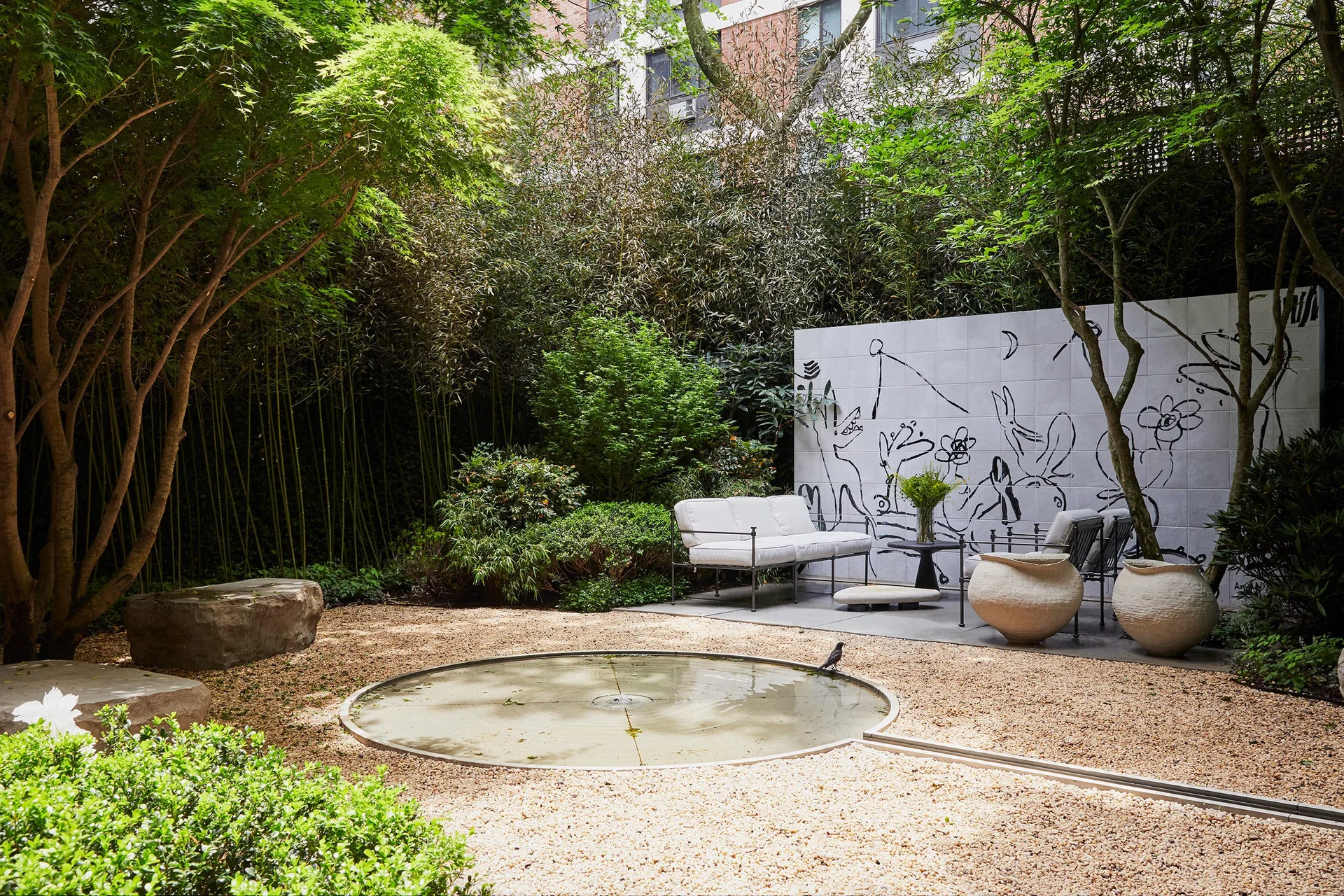 A landscaped garden courtyard features a central circular pond in a gravel yard, a white mural wall with black drawings, and a seating area.
