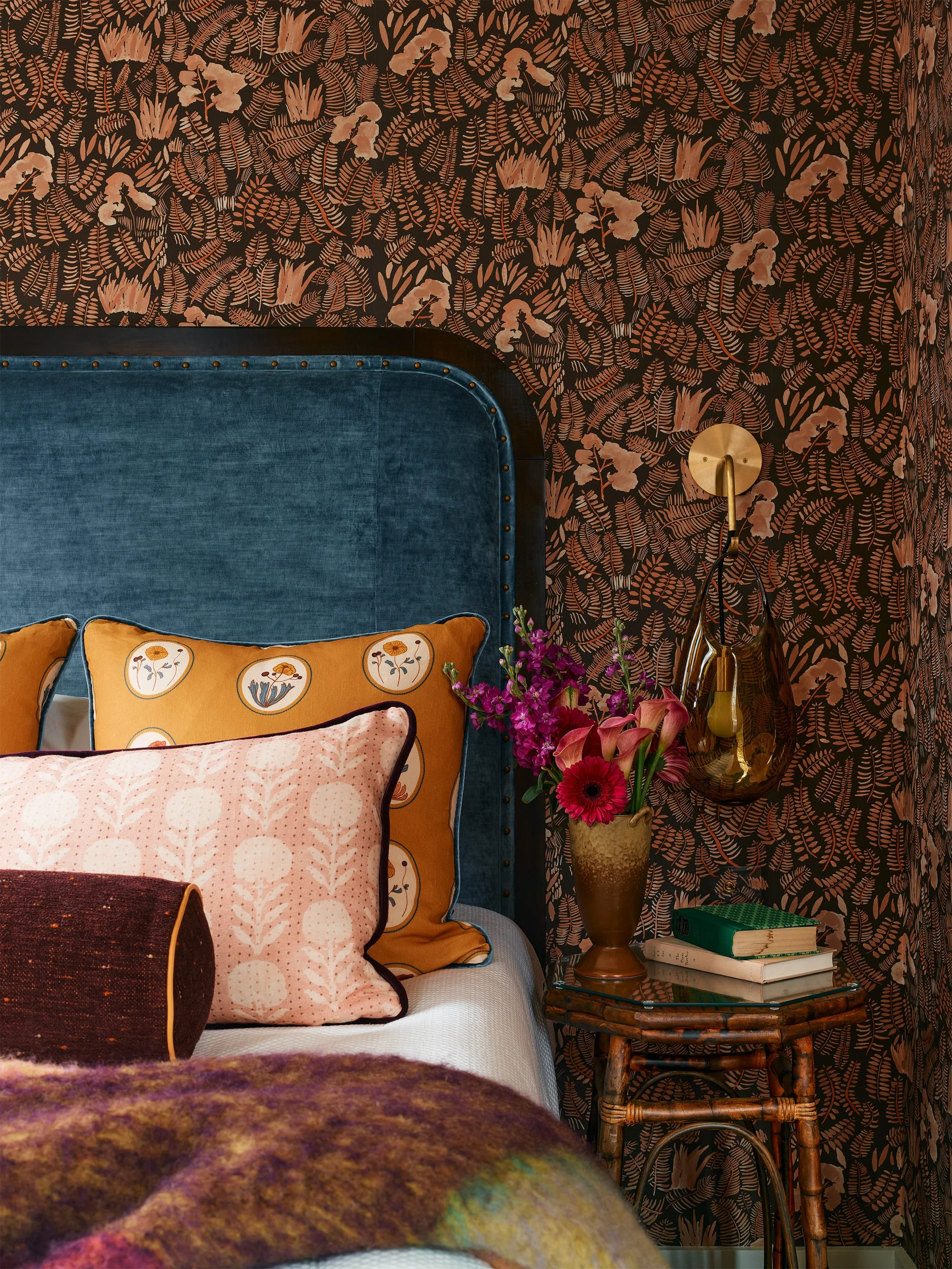 A blue velvet headboard with nailhead trim is styled next to a bamboo nightstand against brown botanical wallpaper in a bedroom.