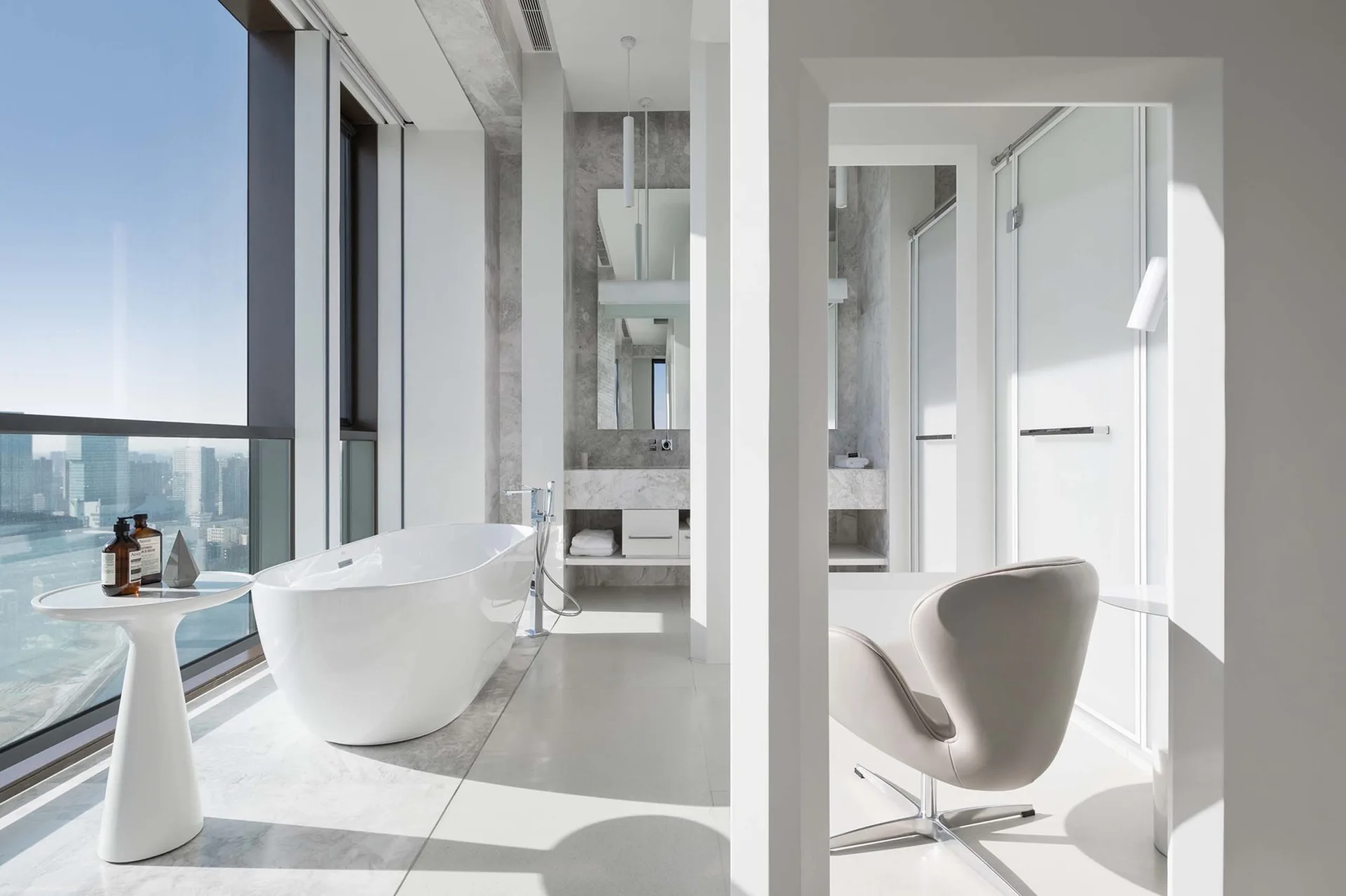 A modern white bathroom featuring a freestanding tub next to large windows, a stone vanity, and a light grey swivel chair.