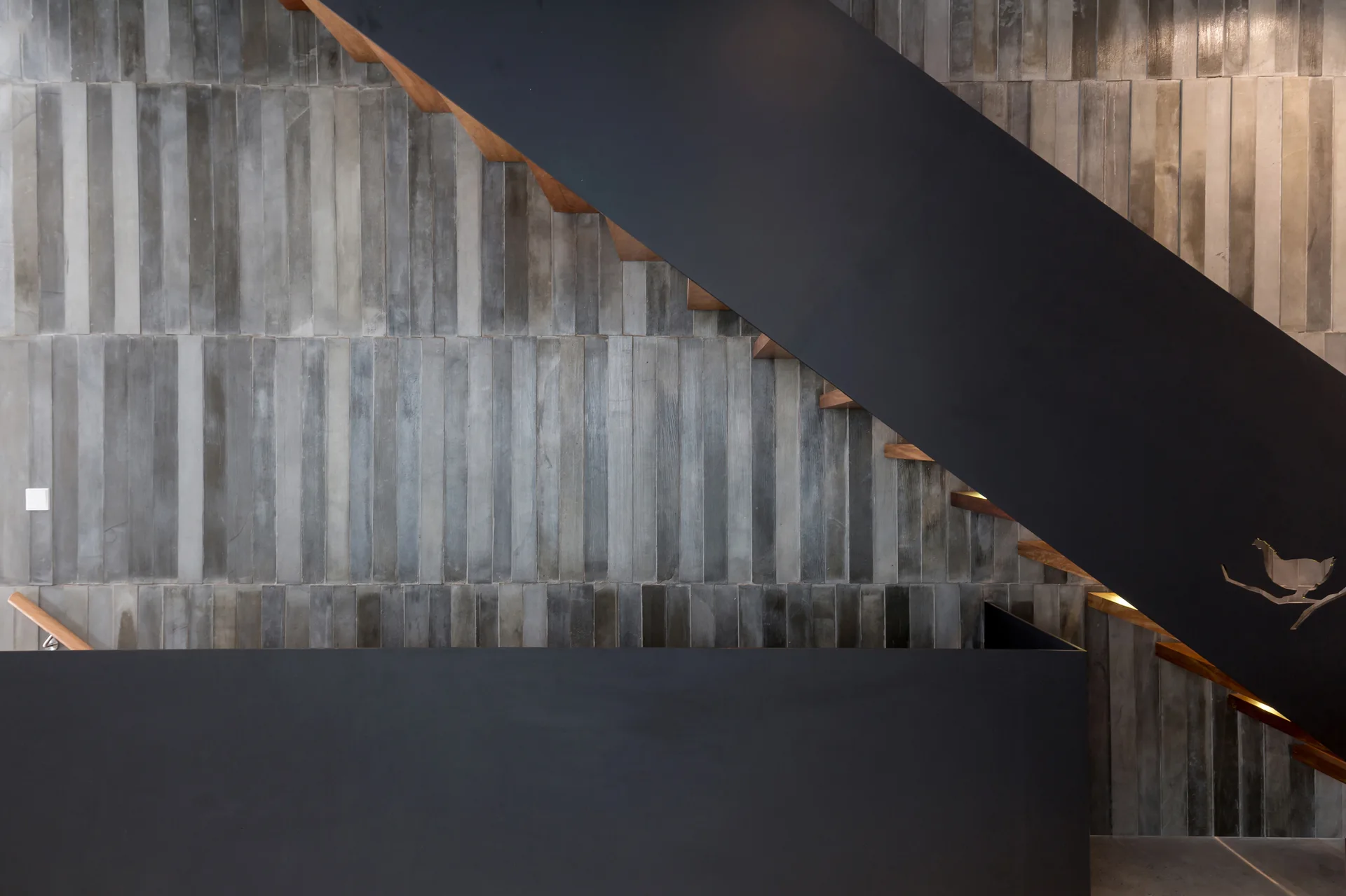 Modern floating wood stairs are mounted against a grey textured tile wall with thick matte black metal plates serving as handrails and guards.