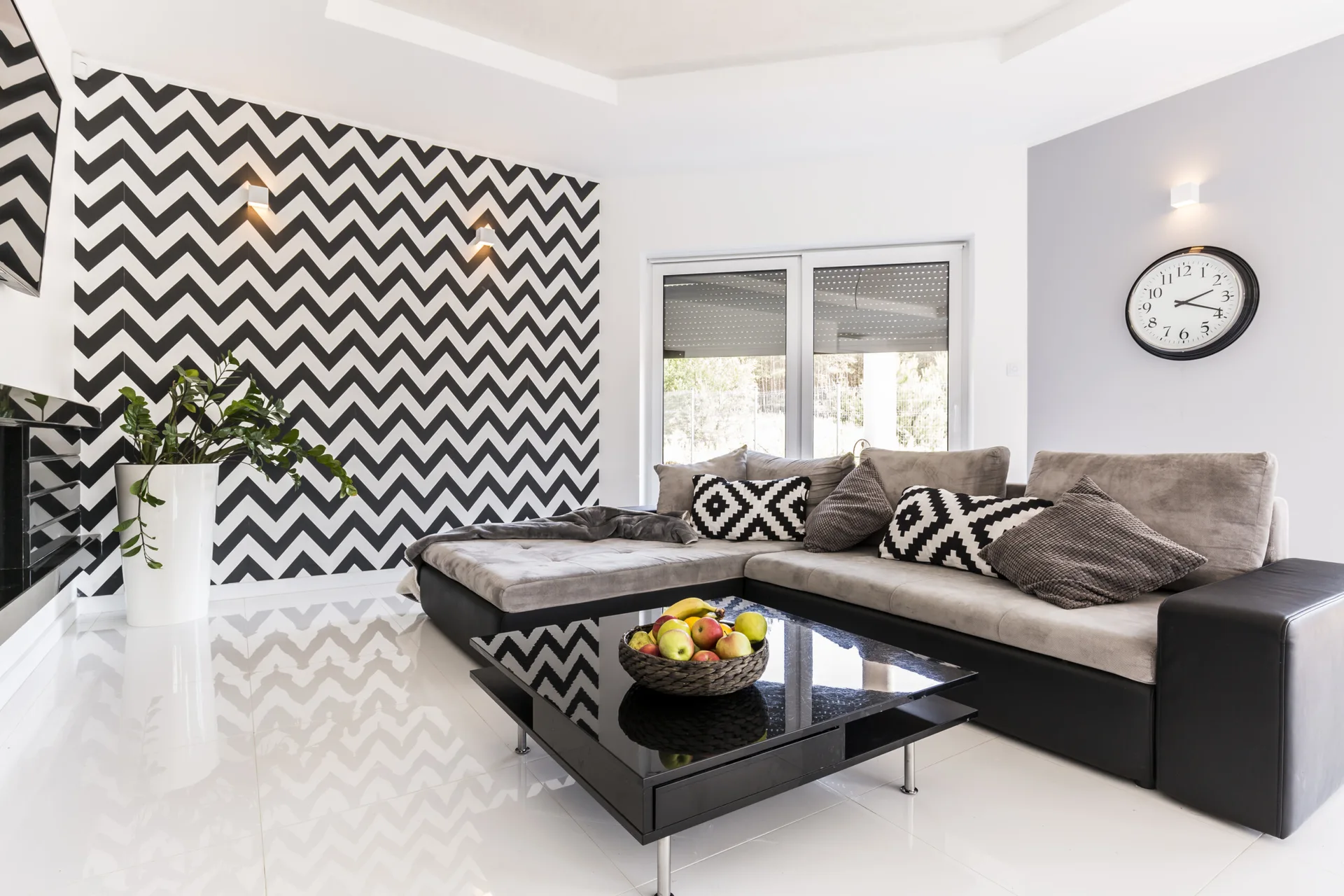 A modern living room featuring a grey sectional sofa, a black glass coffee table, and a bold black and white chevron accent wall.