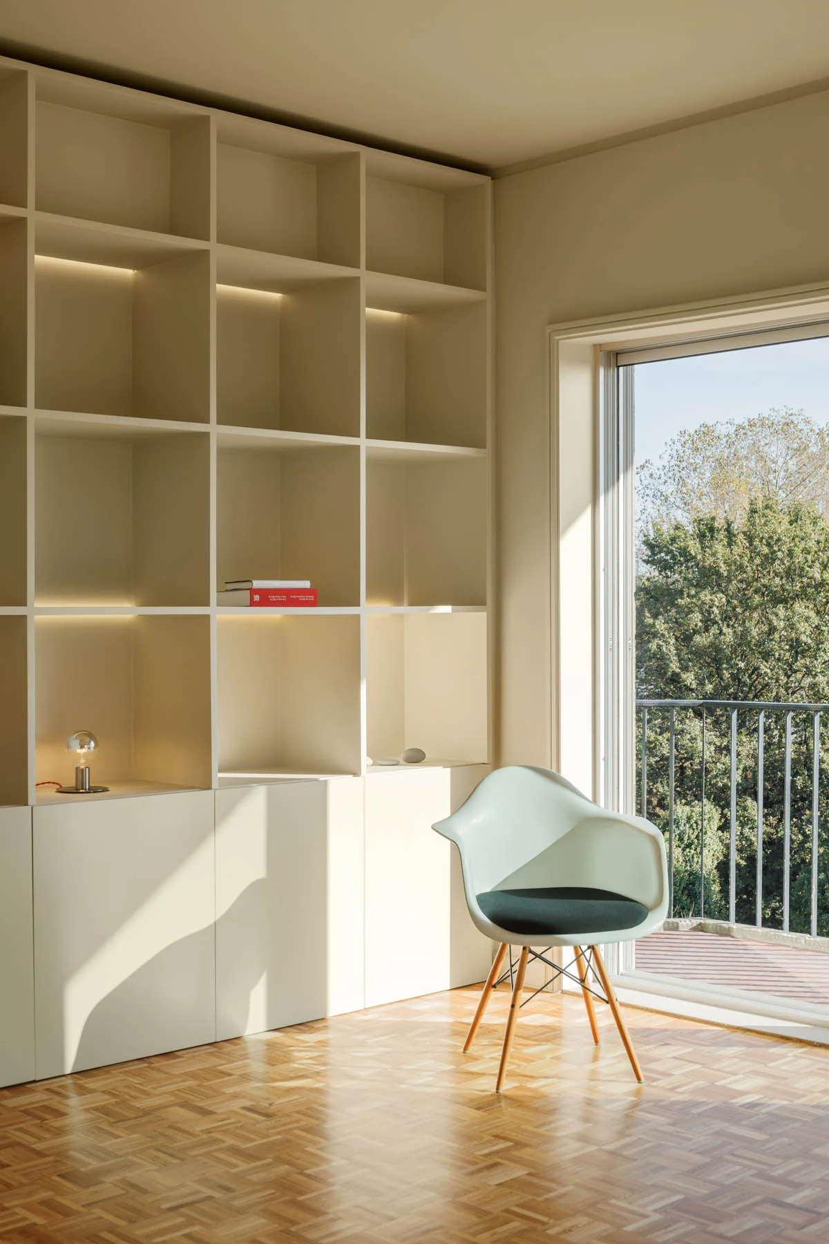 Floor-to-ceiling cream shelving stands next to a white molded armchair on parquet wood flooring by a glass balcony door.