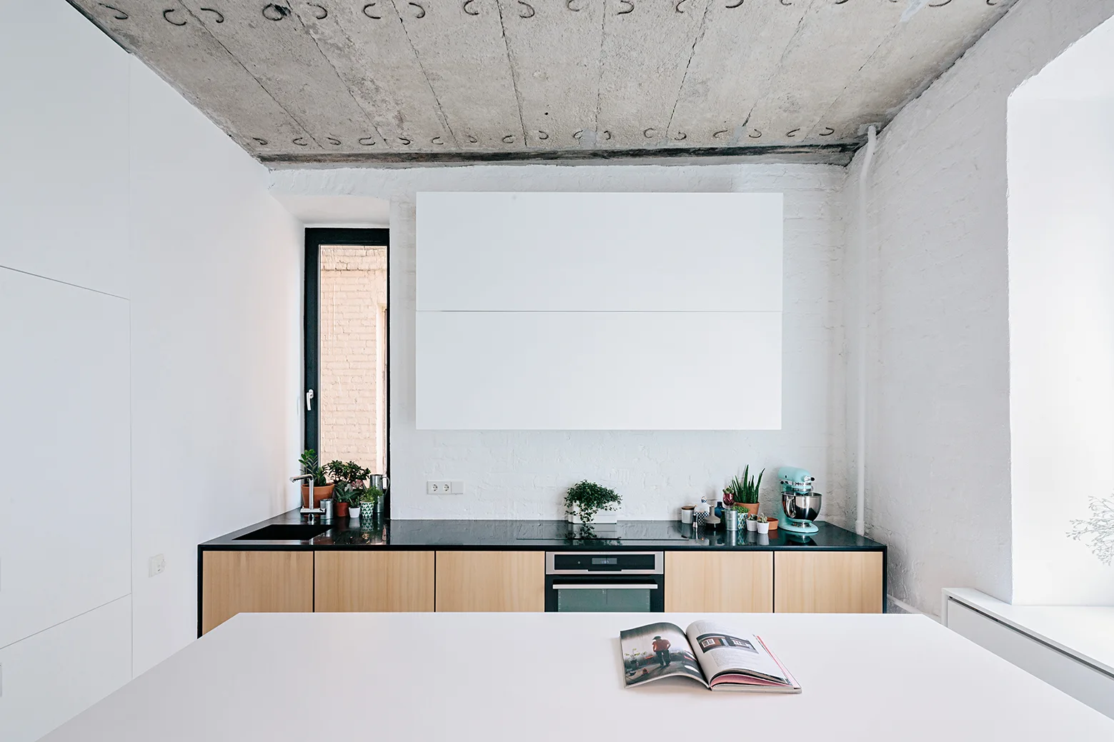 A modern kitchen featuring light wood base cabinets, a black countertop, an exposed concrete ceiling, and white painted brick walls.