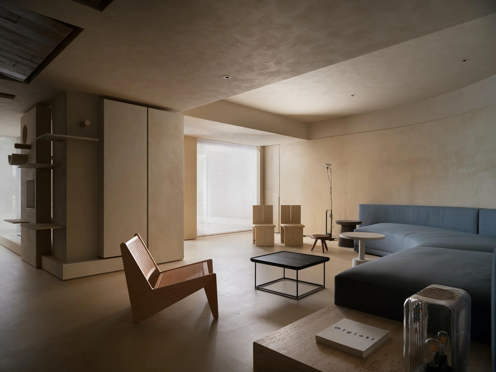 A minimalist beige plaster living room features a blue curved sectional sofa, a wood and cane lounge chair, and sculptural built-in shelving.