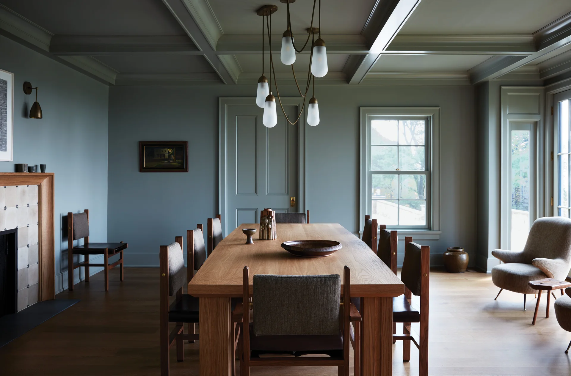 A formal dining room features a long oak table and chairs under a brass chandelier with walls and a coffered ceiling painted in muted blue-grey.