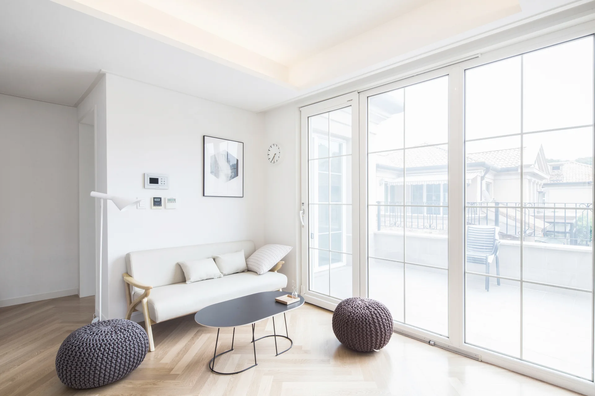 A bright minimalist living room features a cream sofa, an oval black coffee table, two grey knit poufs, and large glass patio doors.