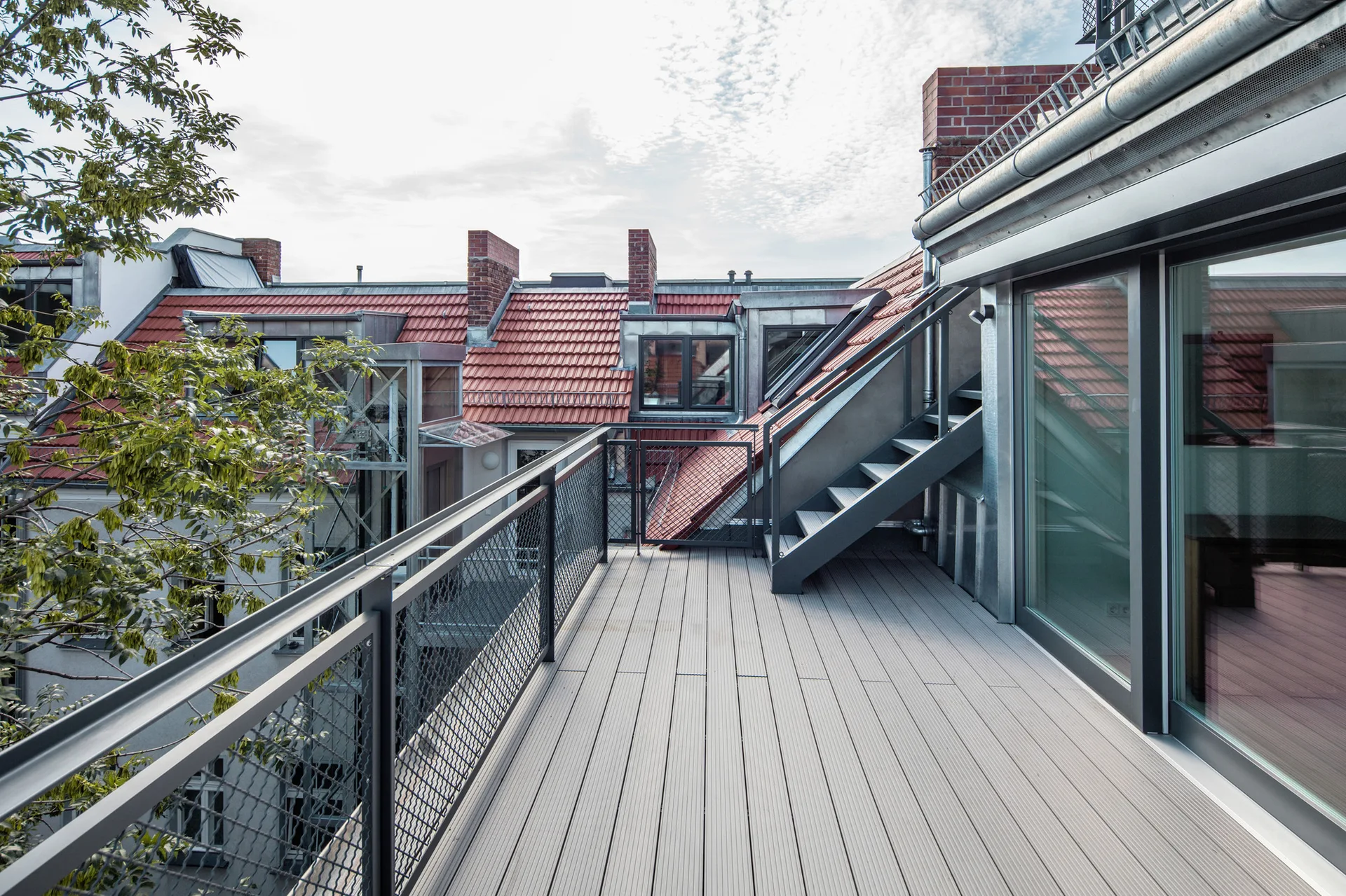 An elevated rooftop balcony features gray composite deck planks, a metal mesh railing, and steel stairs leading up against a background of red roof tiles.