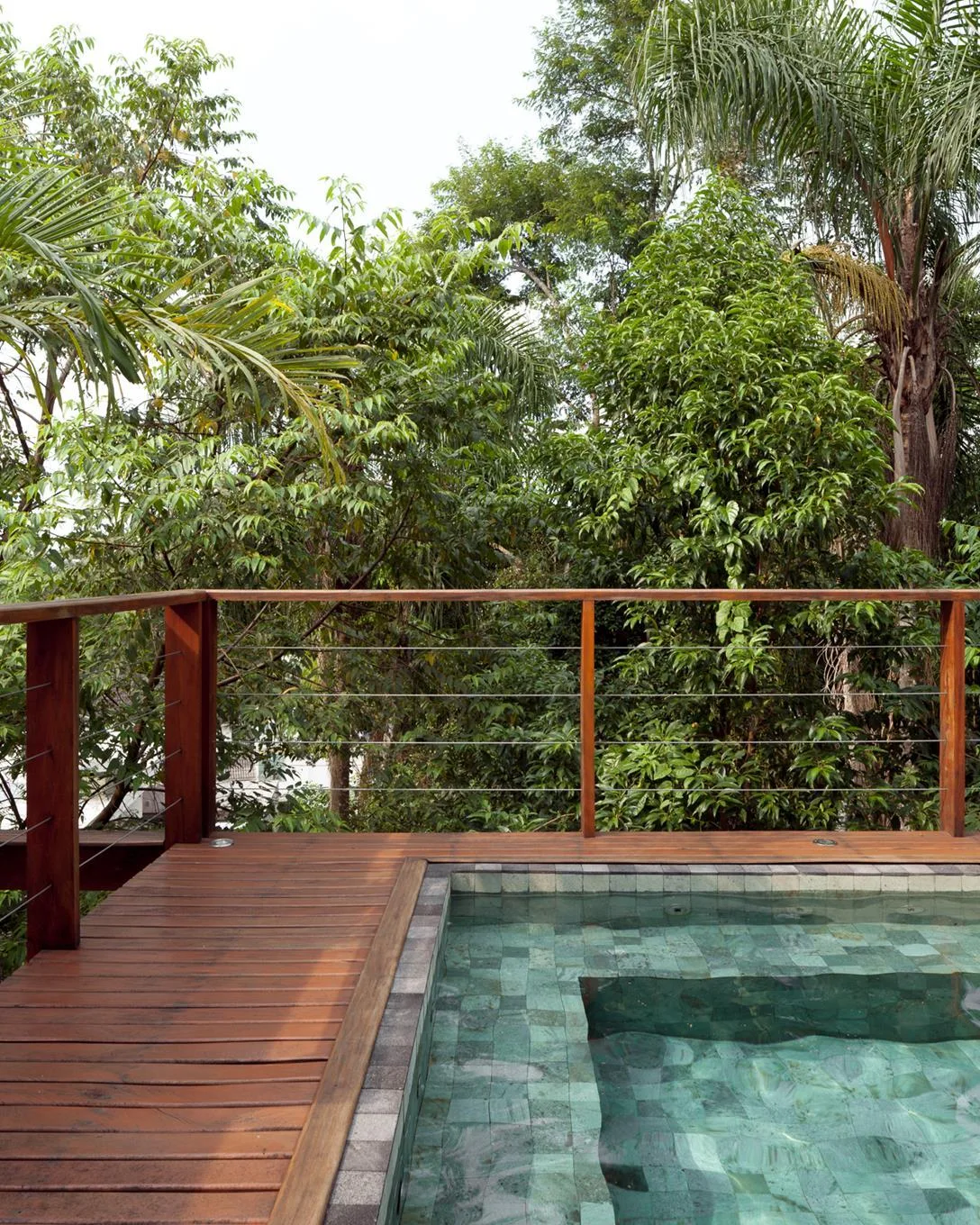 An elevated wooden pool deck with a cable railing overlooks dense green trees, showcasing a swimming pool lined with green tiles.