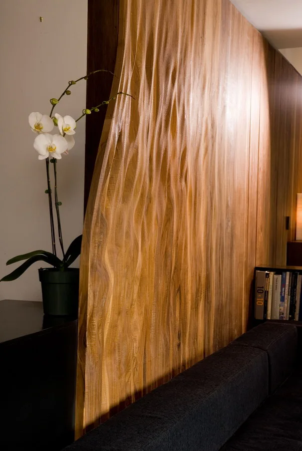 A textured wood partition wall with wavy vertical ridges stands behind a dark sofa next to a white orchid in a pot.