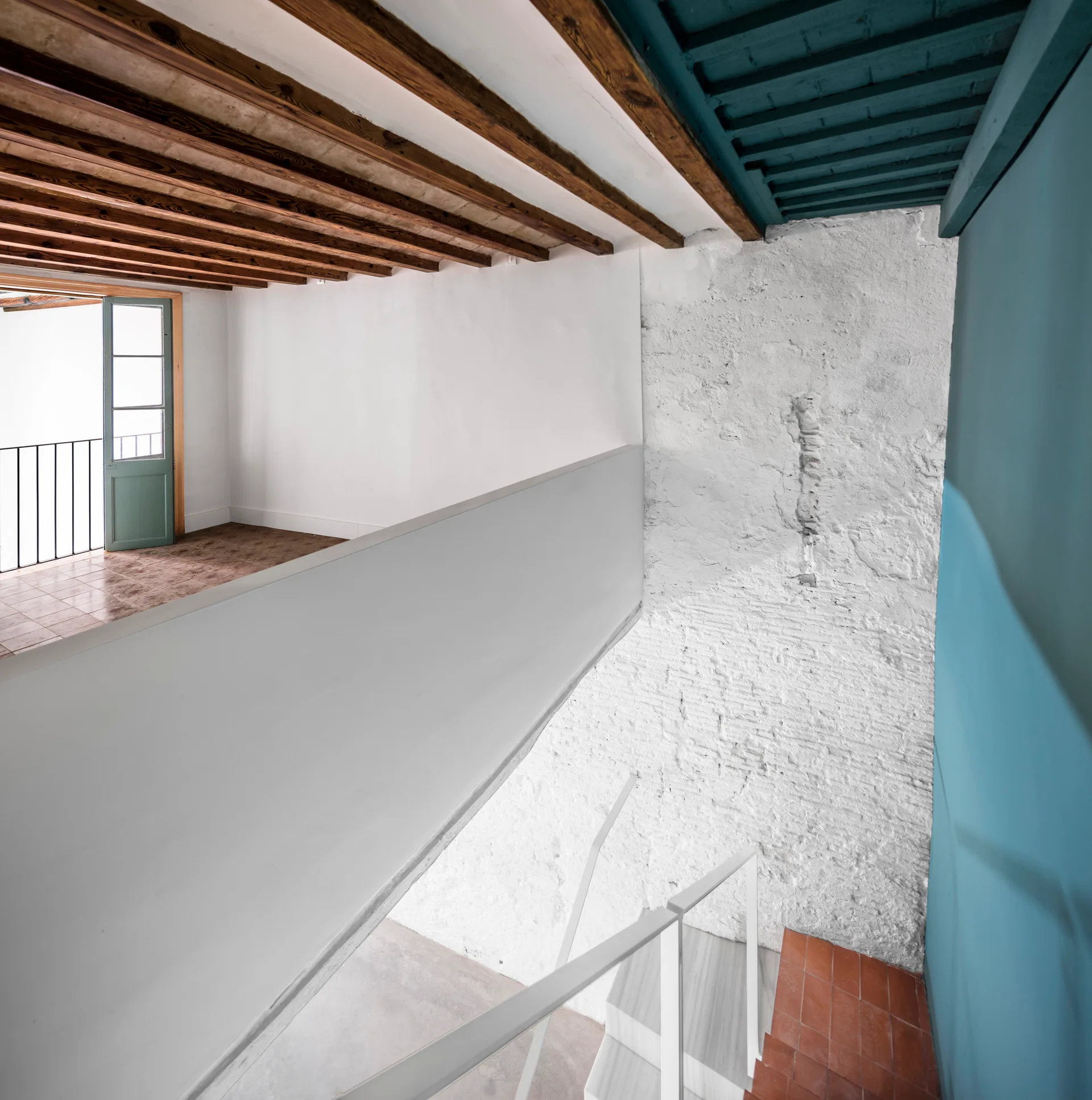 A minimalist loft staircase features a white metal railing, an exposed wooden beam ceiling, a white painted brick wall, and a blue painted accent wall.