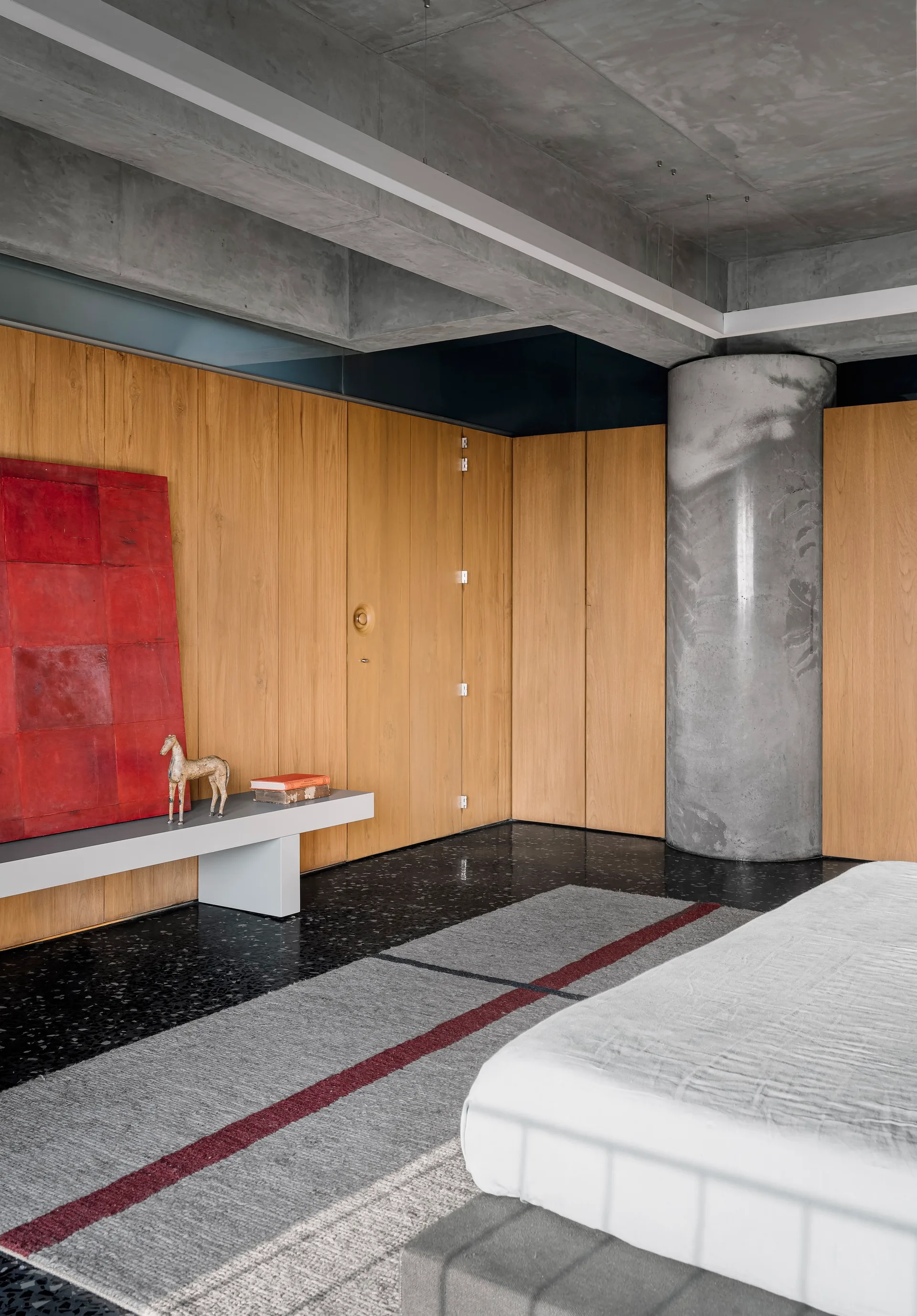 A minimalist bedroom featuring wood-paneled walls, a large raw concrete column, and a dark speckled terrazzo floor next to a platform bed.