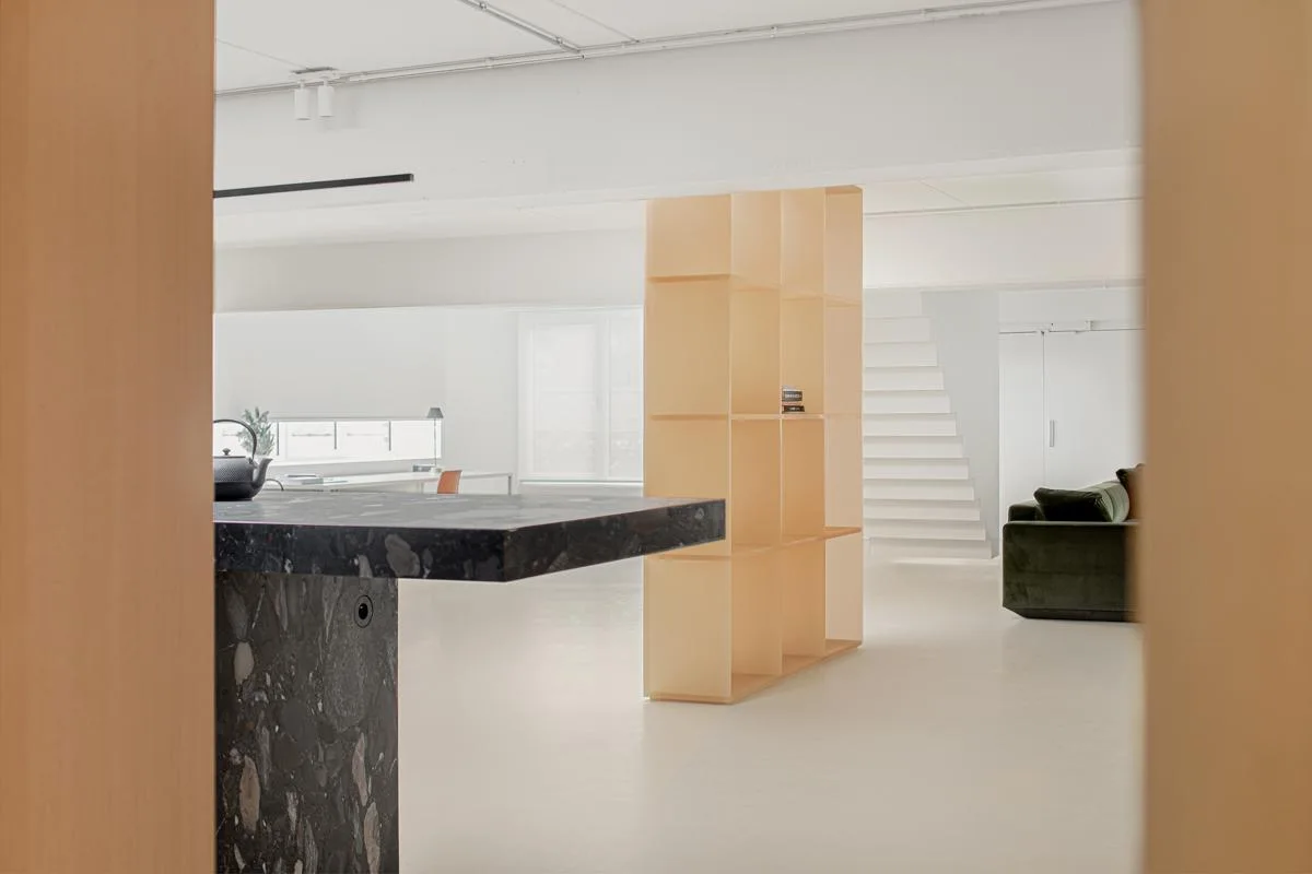 A dark stone kitchen island stands in a minimalist white open-plan space with a light wood partition bookshelf and a white staircase in the background.
