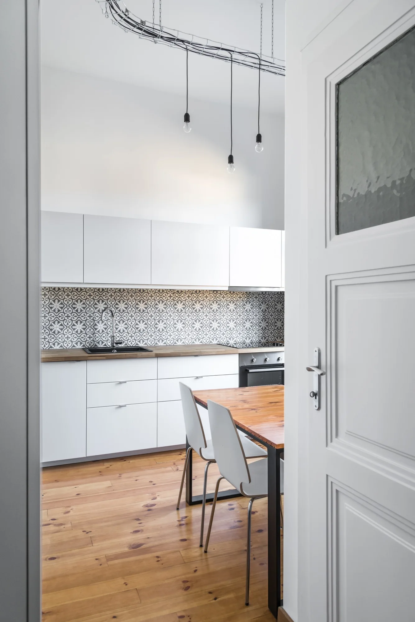 A white door opens to reveal a kitchen featuring flat-front white cabinets, a black and white patterned tile backsplash, and a wooden dining table.