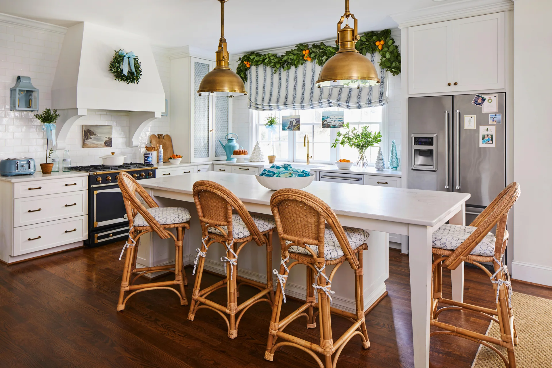 A bright white kitchen featuring a large central island with four rattan bar stools, two brass pendant lights, and a window with a striped shade.
